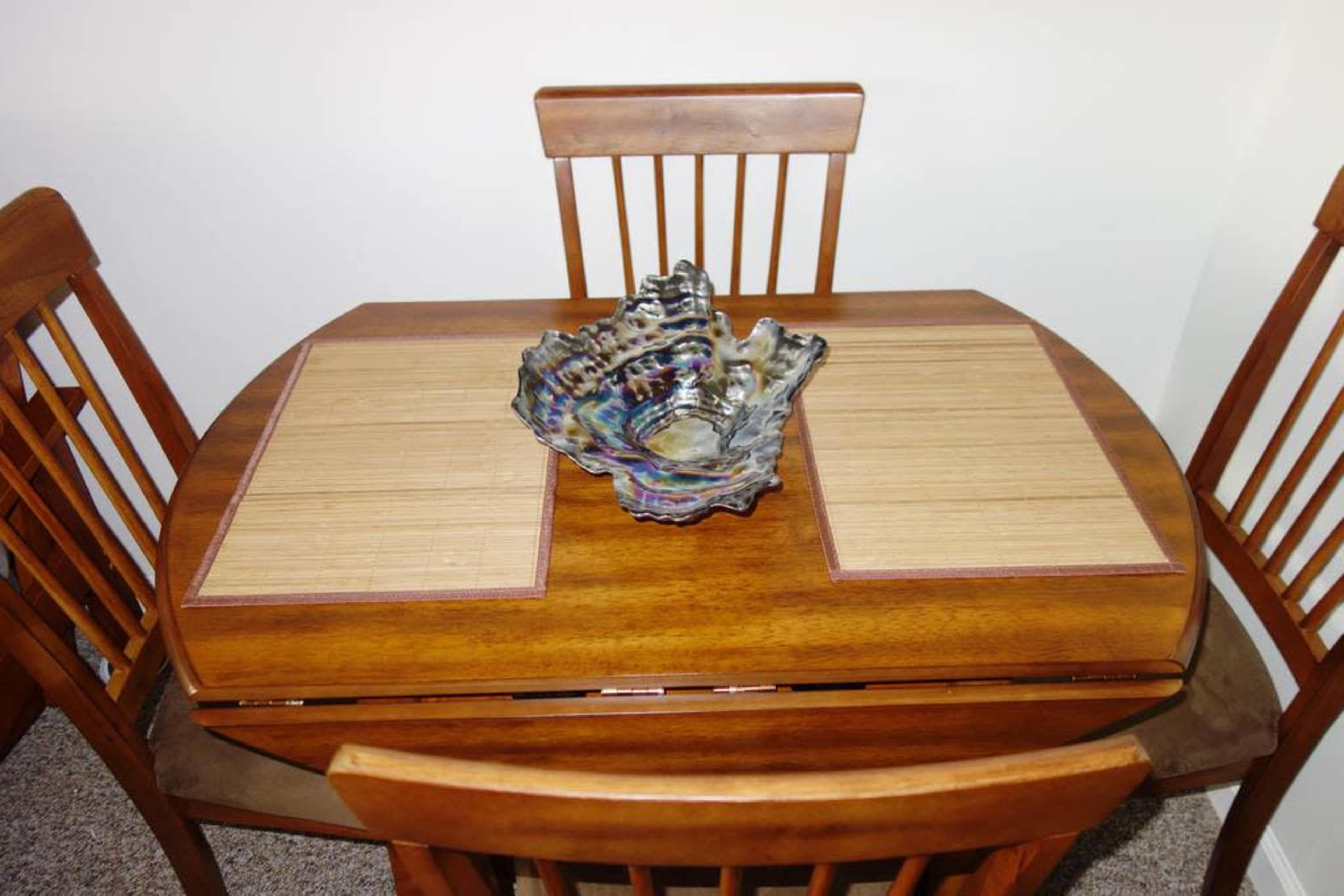 A wooden dining table with bamboo placemats is set with a decorative glass bowl in the center, surrounded by four wooden chairs.