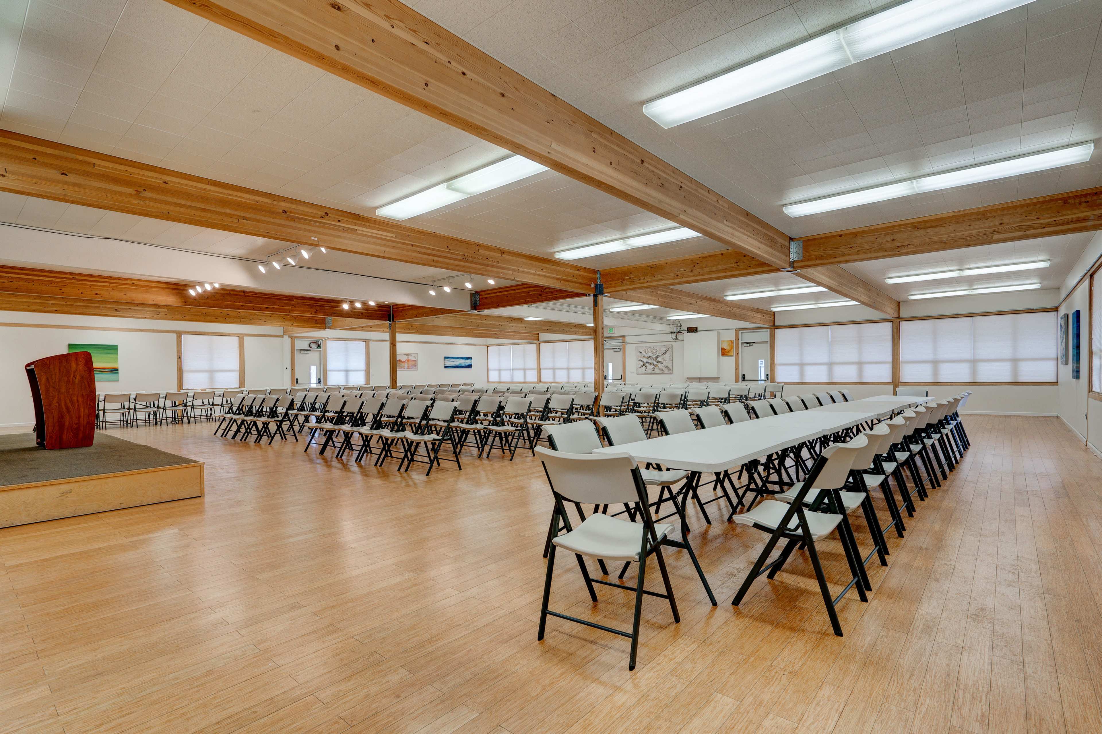 A spacious indoor hall with rows of folding tables and chairs arranged for an event, featuring wooden beams in the ceiling and a small stage area.
