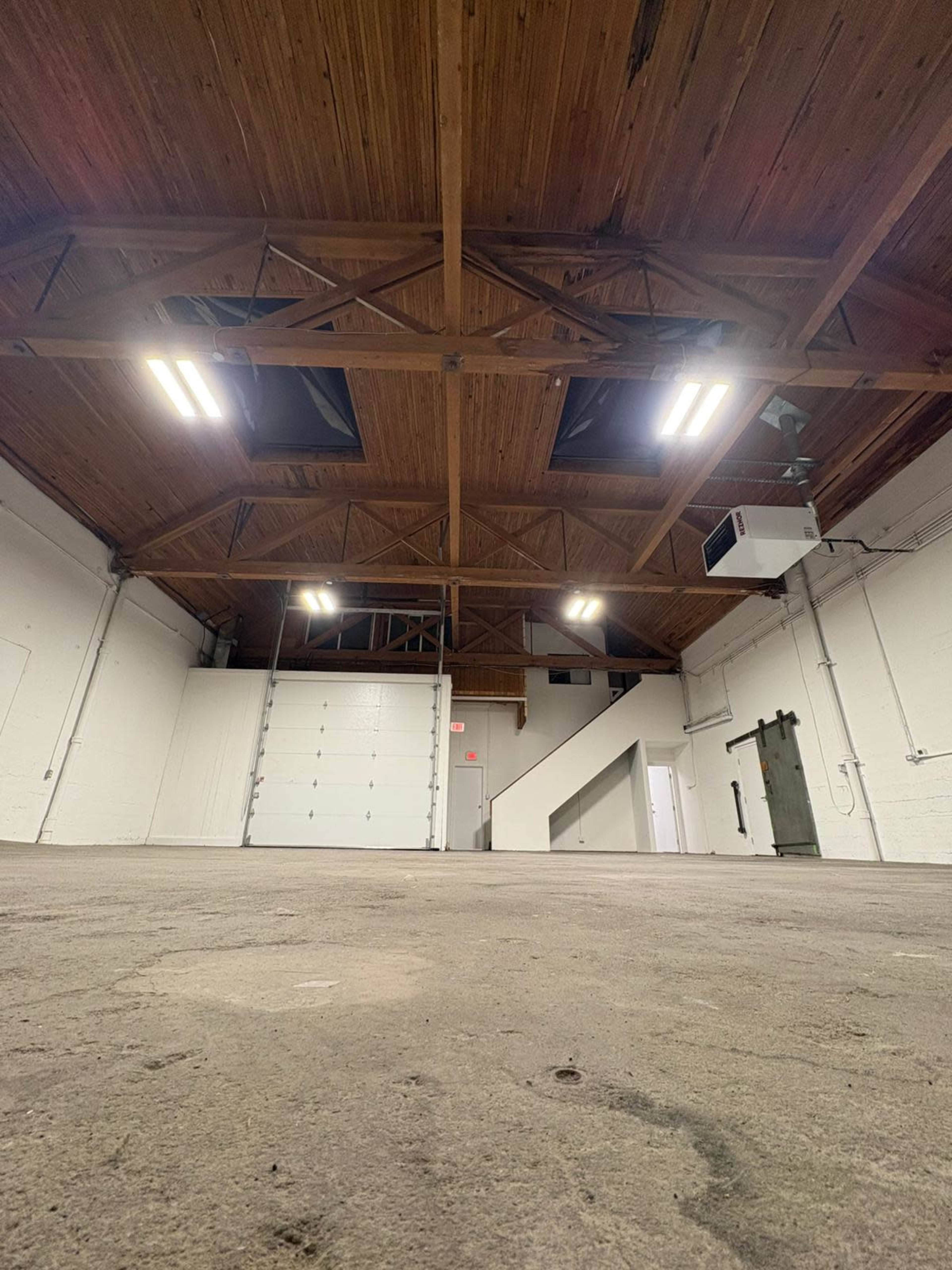 The image shows a spacious, empty warehouse interior with a high wooden ceiling, large overhead lights, and a roll-up door.