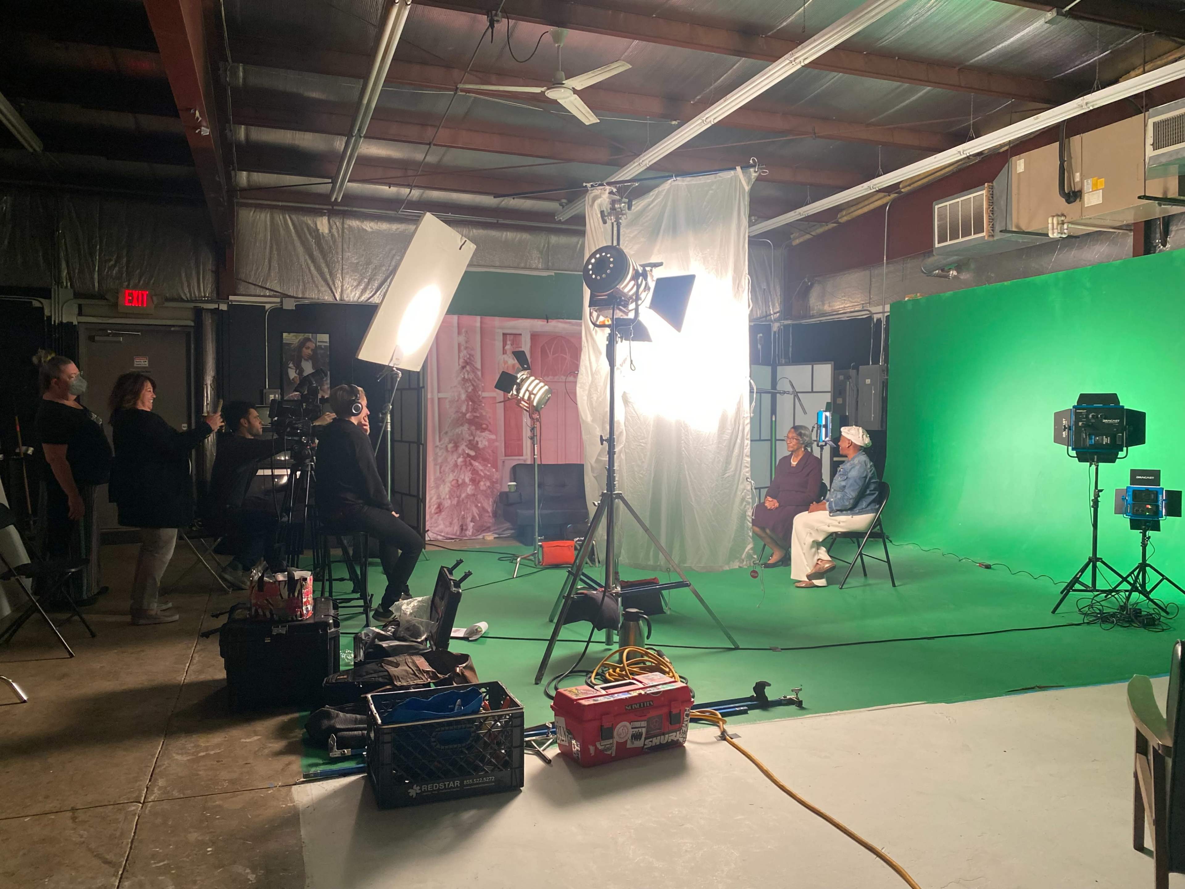 A film crew is set up in a studio with green screen backgrounds, lights, and cameras, preparing to record a scene with two seated individuals.