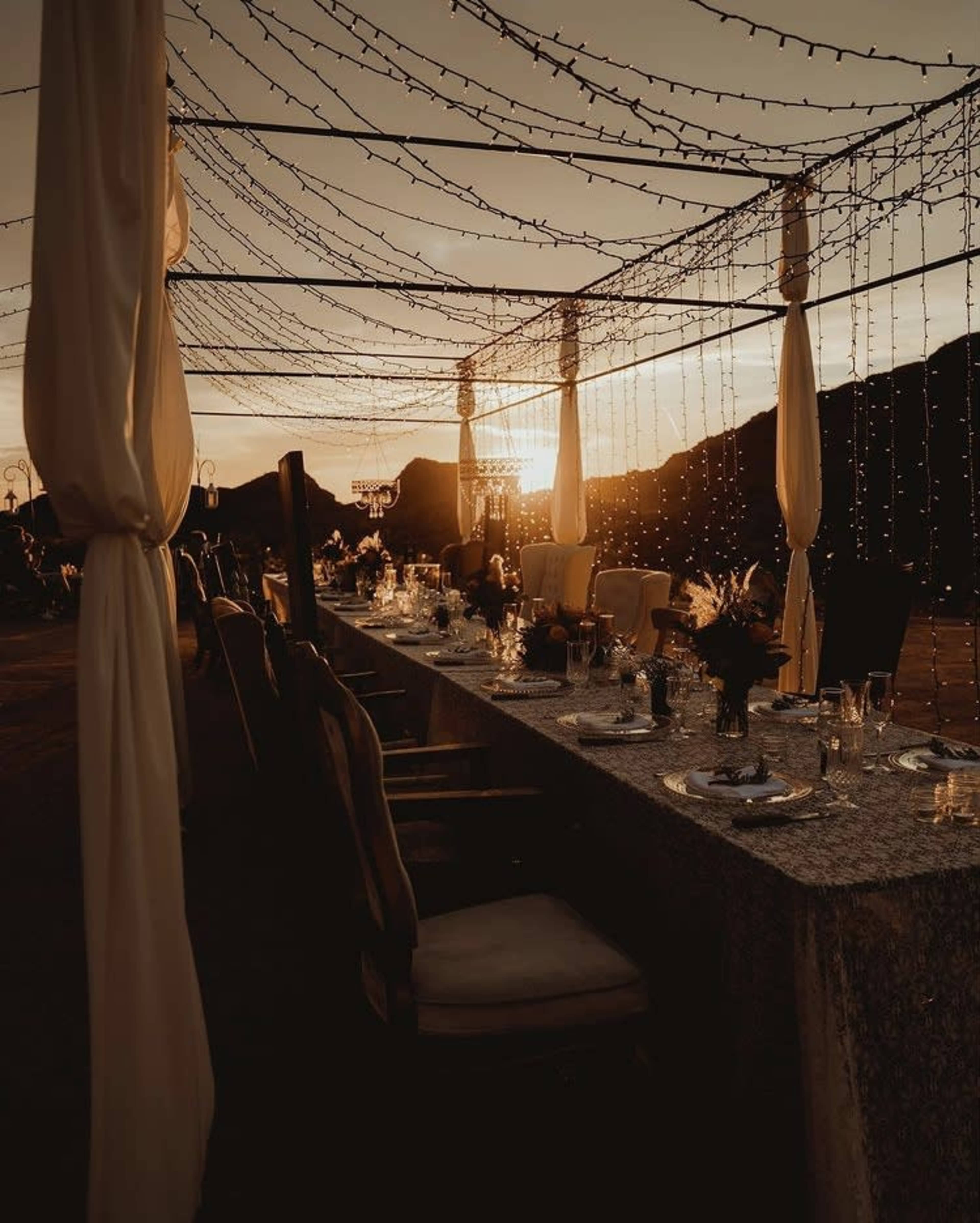 A long table is set for a dinner under string lights at sunset, with mountains in the background.