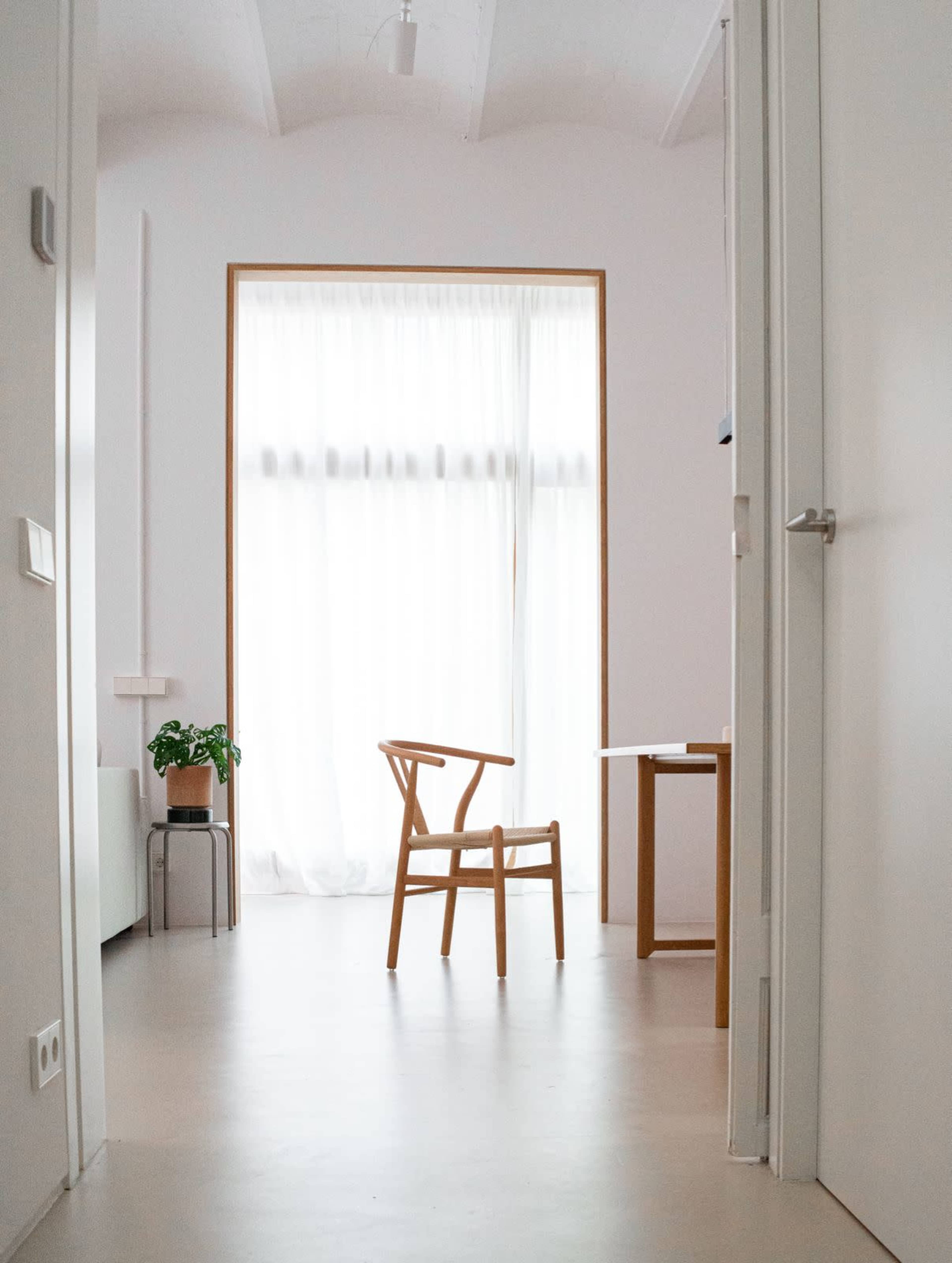The image shows a minimalist interior space with a wooden chair facing a window covered by sheer curtains, and a small potted plant nearby.