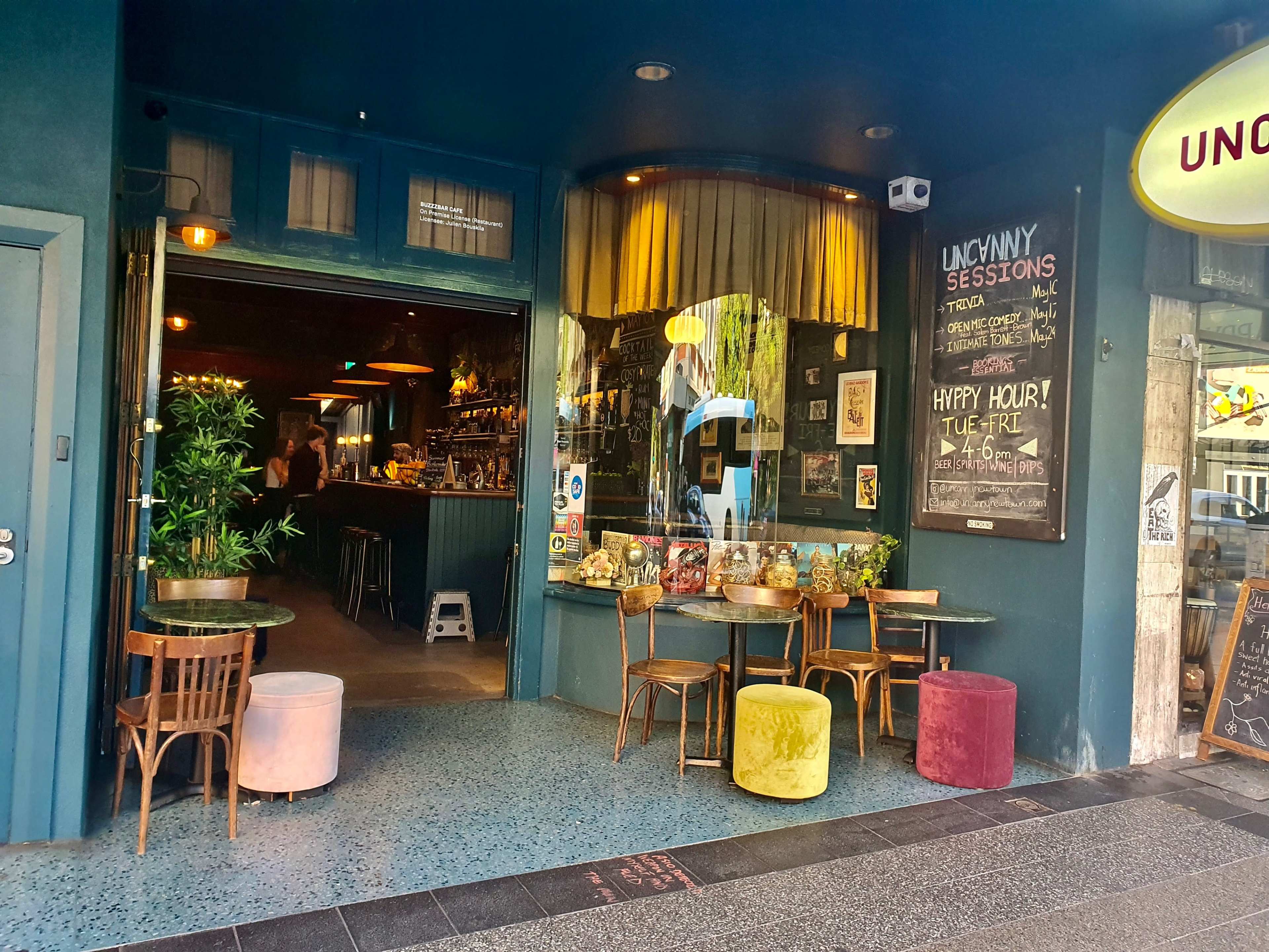The entrance of a bar with colorful stools and a chalkboard menu, featuring indoor seating and plants visible through the open doorway.