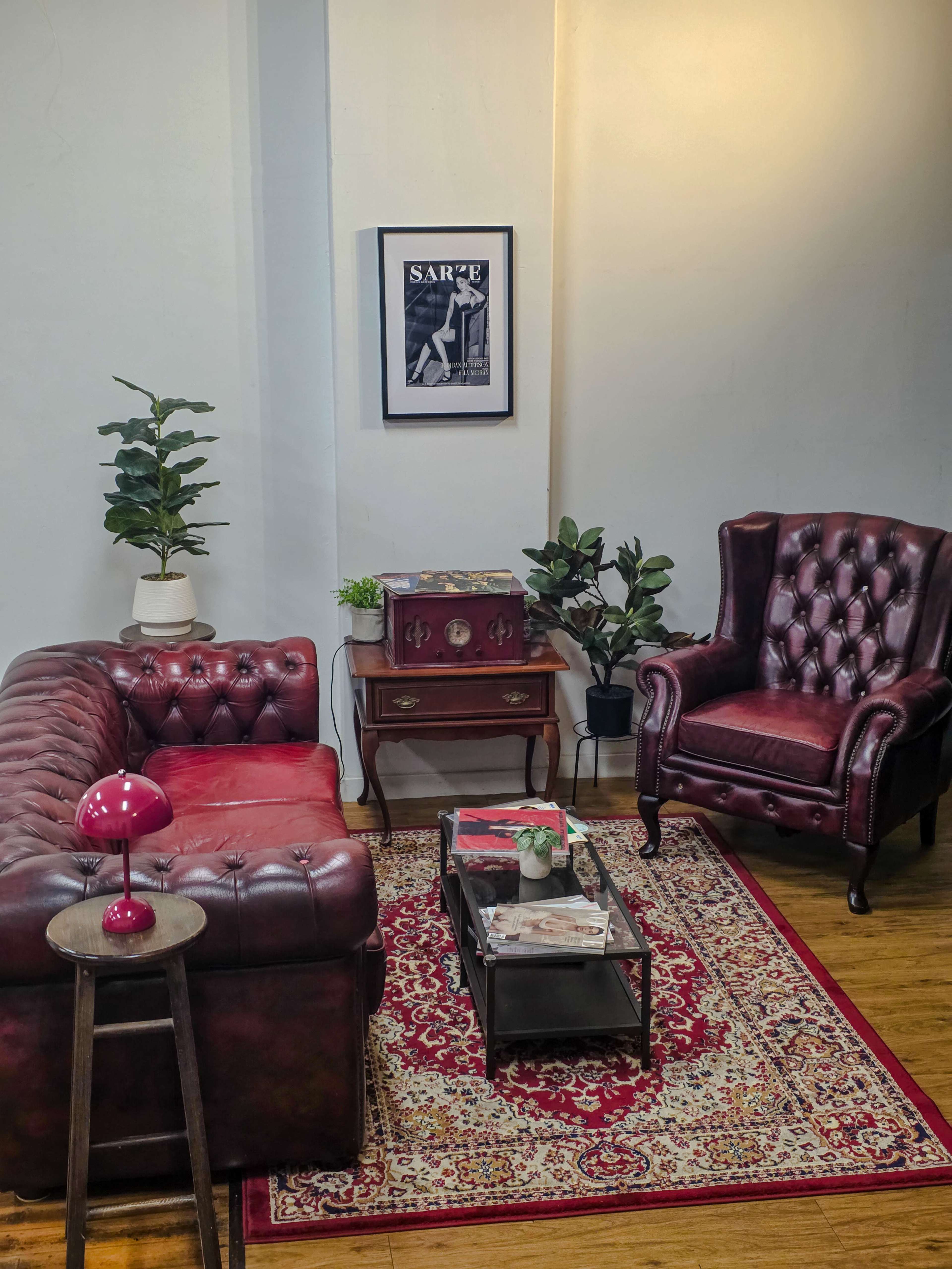 The image features a cozy living room setup with a chesterfield sofa and a wingback chair, complemented by a coffee table and decorative plants against a light wall.