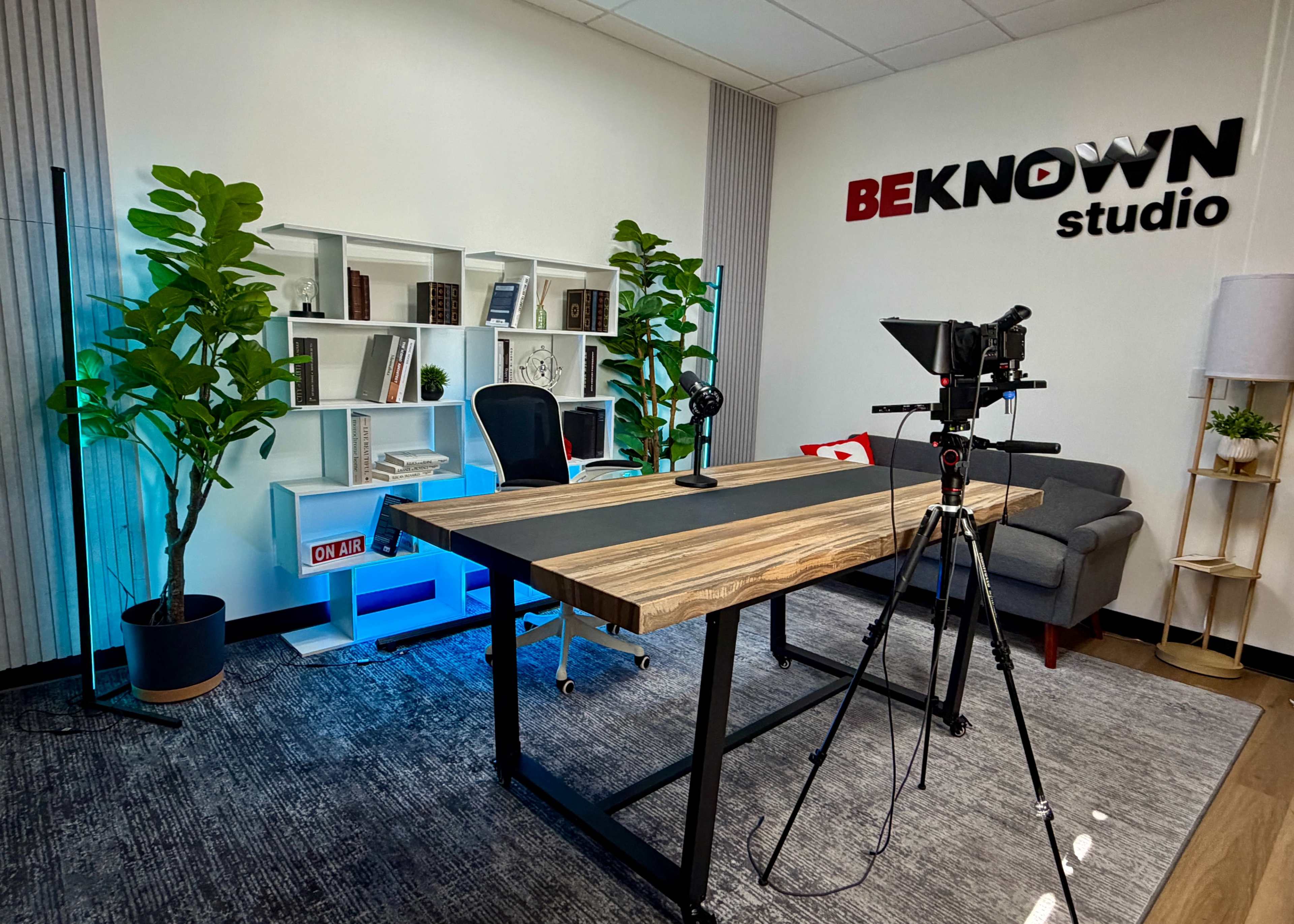 The image shows a well-organized studio space featuring a large wooden table, a microphone setup, a camera on a tripod, a comfortable gray couch, and a bookshelf filled with books and plants against the wall.