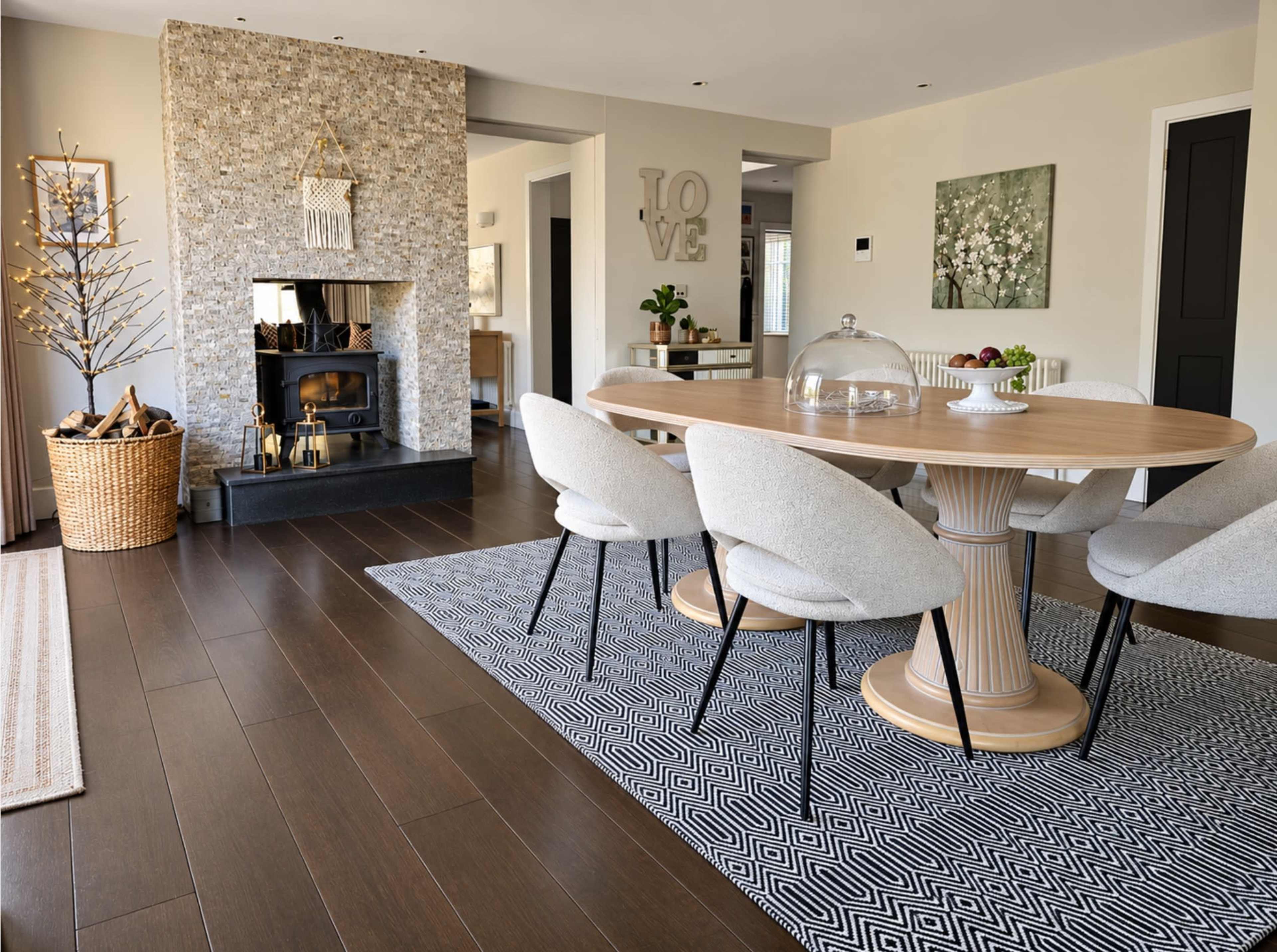 A modern dining area features a round wooden table surrounded by upholstered chairs, set against a textured stone fireplace and a patterned rug.
