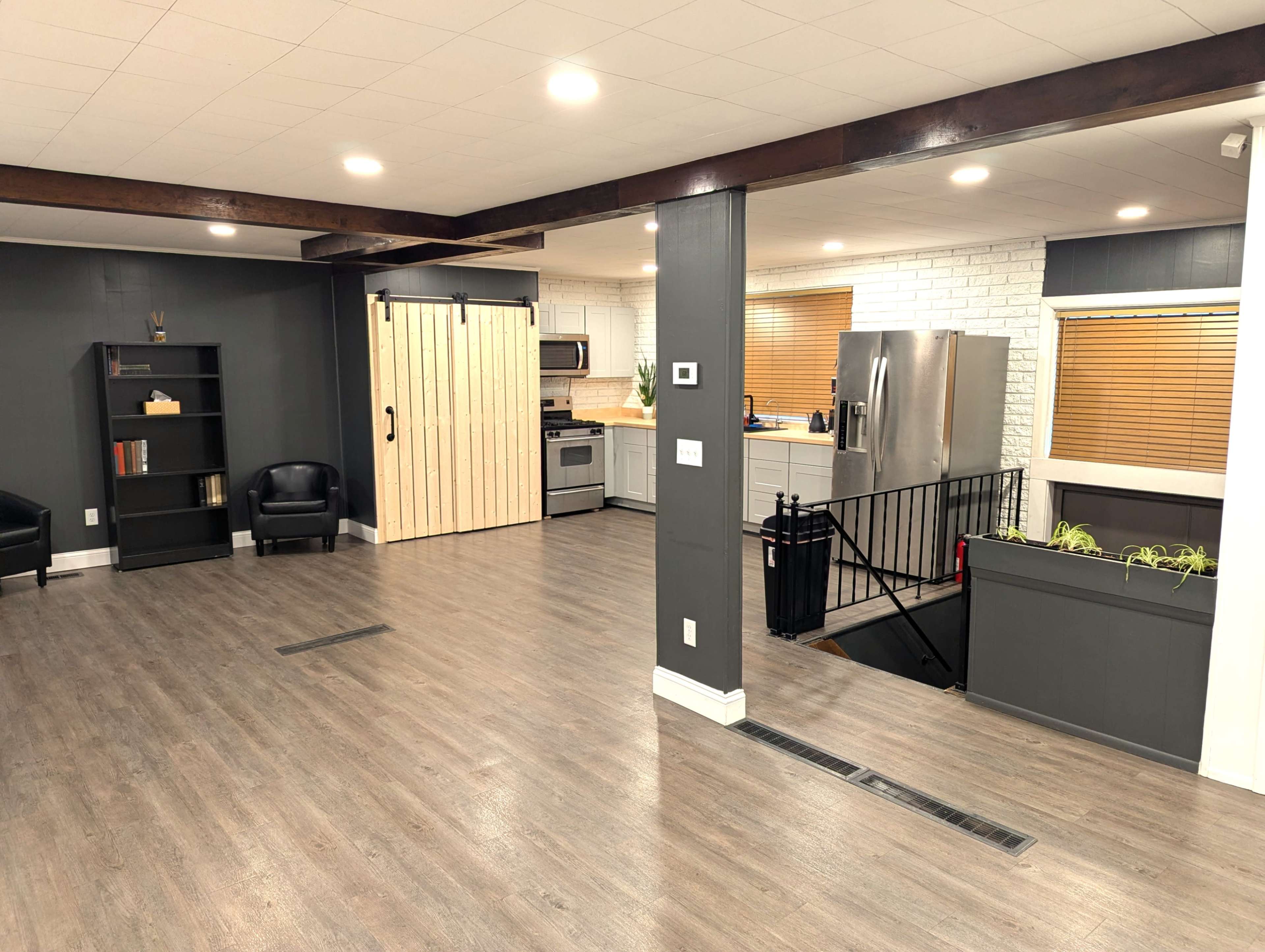 The image shows an open-concept living area with a modern kitchen, a sliding wooden door, a bookshelf, and a staircase leading to a lower level.