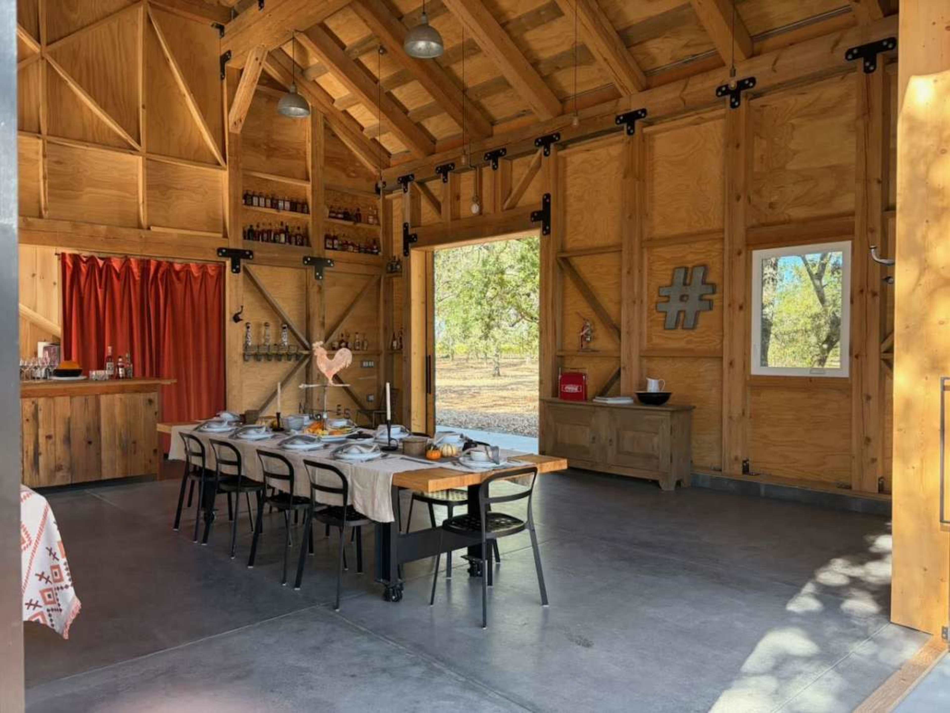 The image shows a rustic dining area inside a wooden structure, featuring a long table set for a meal, with chairs arranged around it and large sliding glass doors revealing an outdoor view.