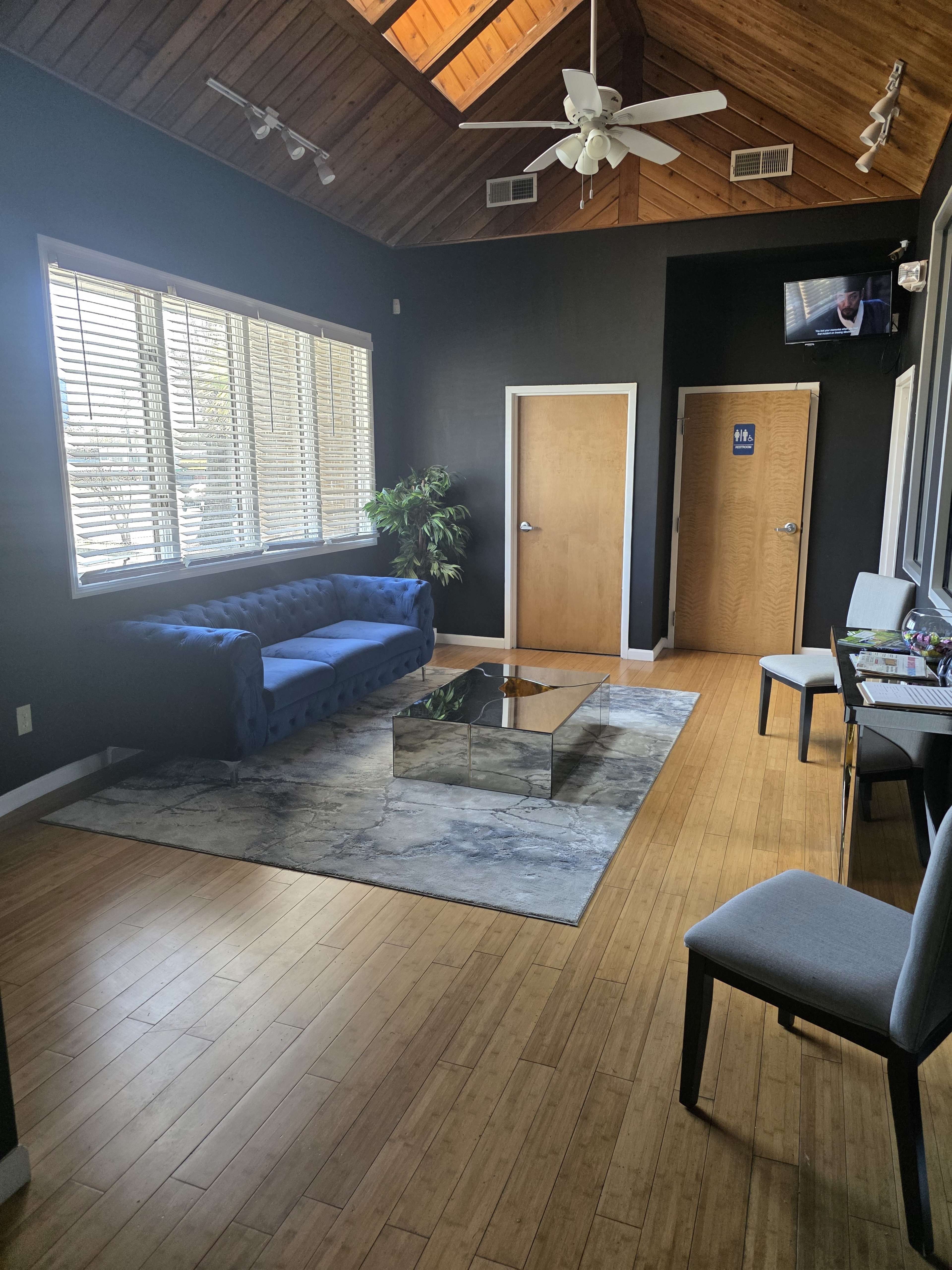 The image shows a waiting room with a blue couch, a glass coffee table, and two doors, all under a wood-paneled ceiling with a ceiling fan.
