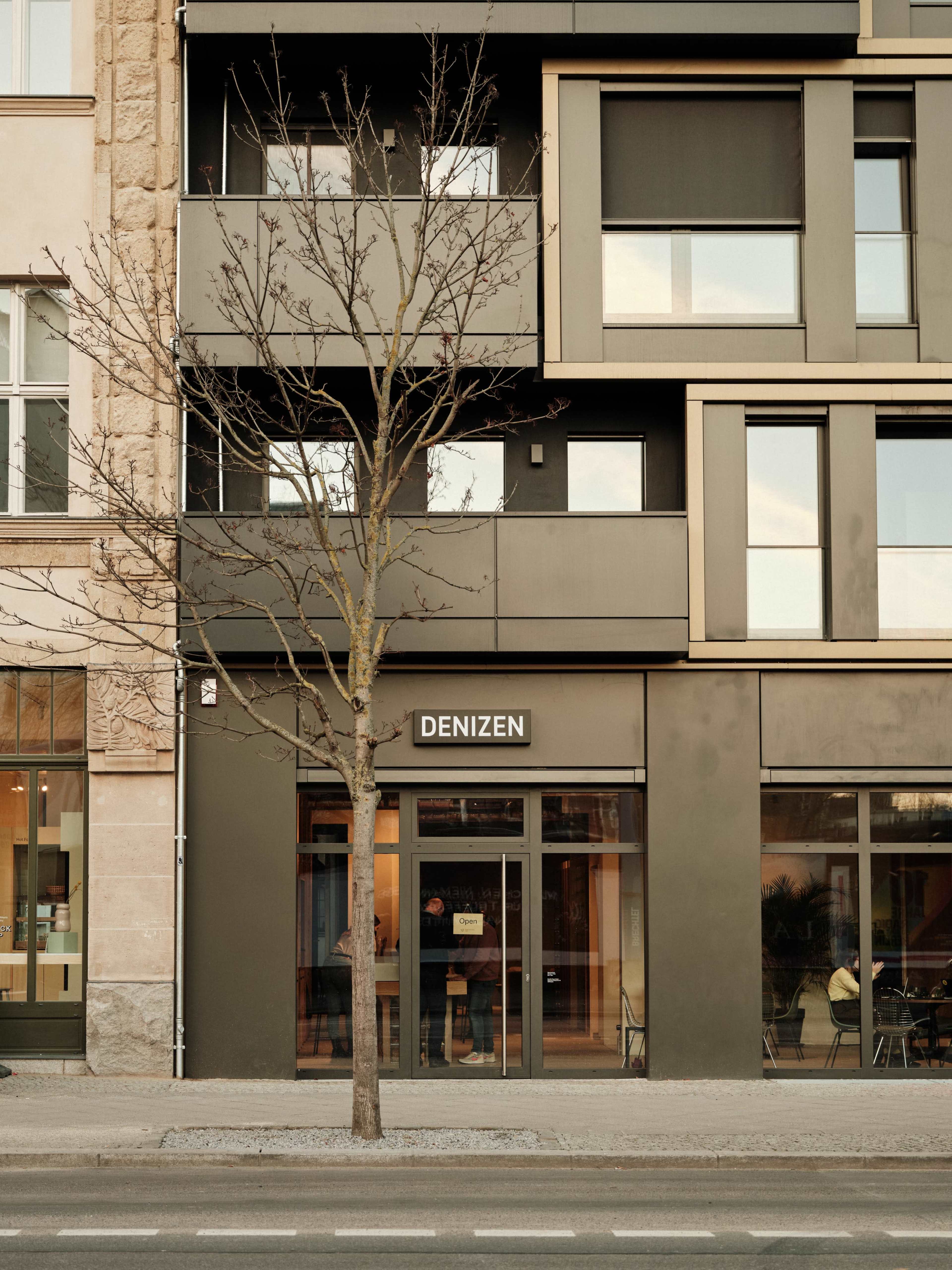 A modern building facade featuring a ground-level storefront labeled "DENIZEN," with a bare tree in front and large windows above.