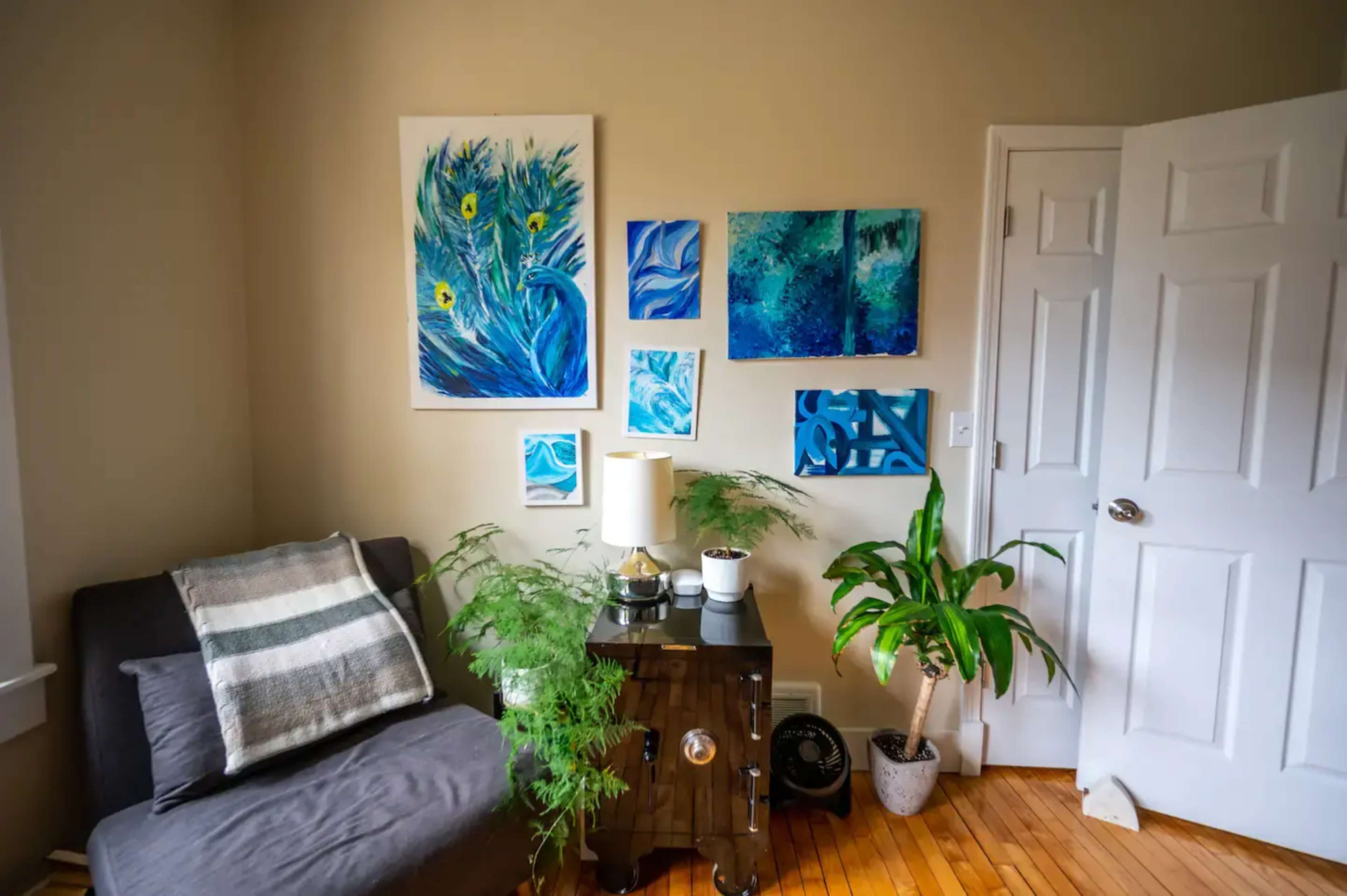 A cozy corner of a room features a grey sofa, various blue paintings on the wall, a leafy plant next to a wooden cabinet, and a white lamp.