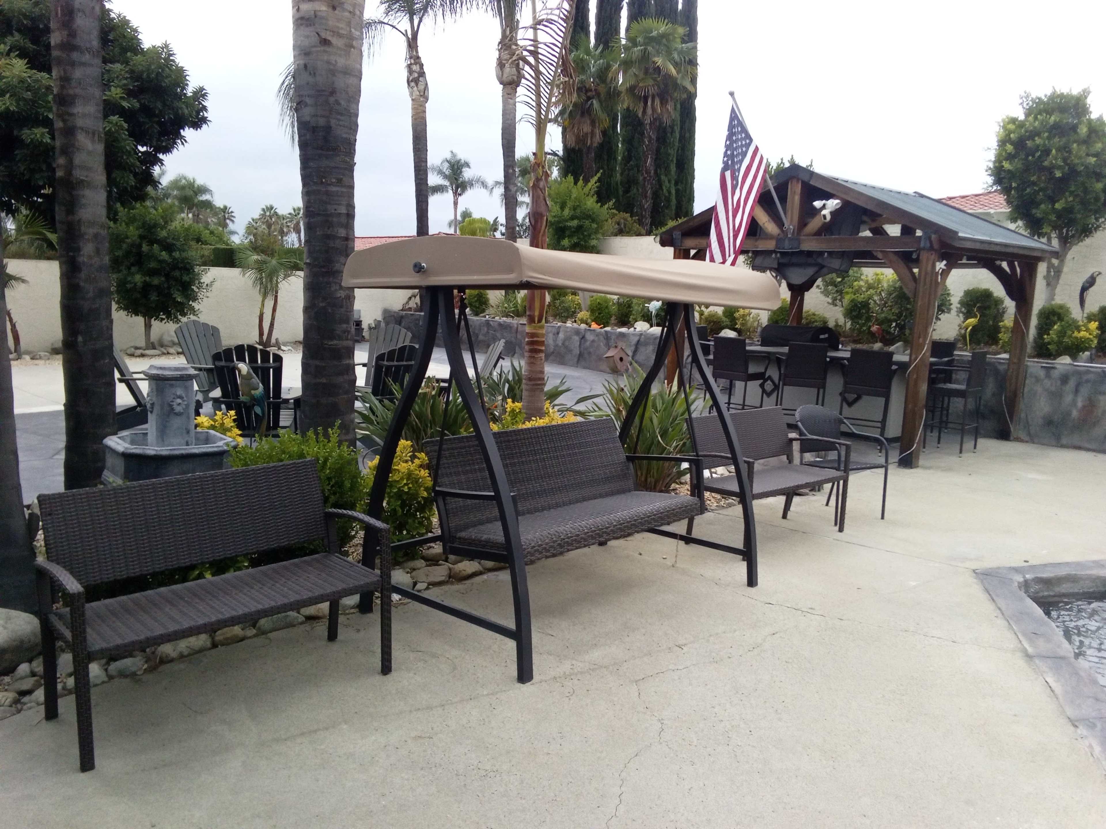 The scene features a patio area with a swing bench, several chairs, and a wooden gazebo, surrounded by palm trees and greenery.