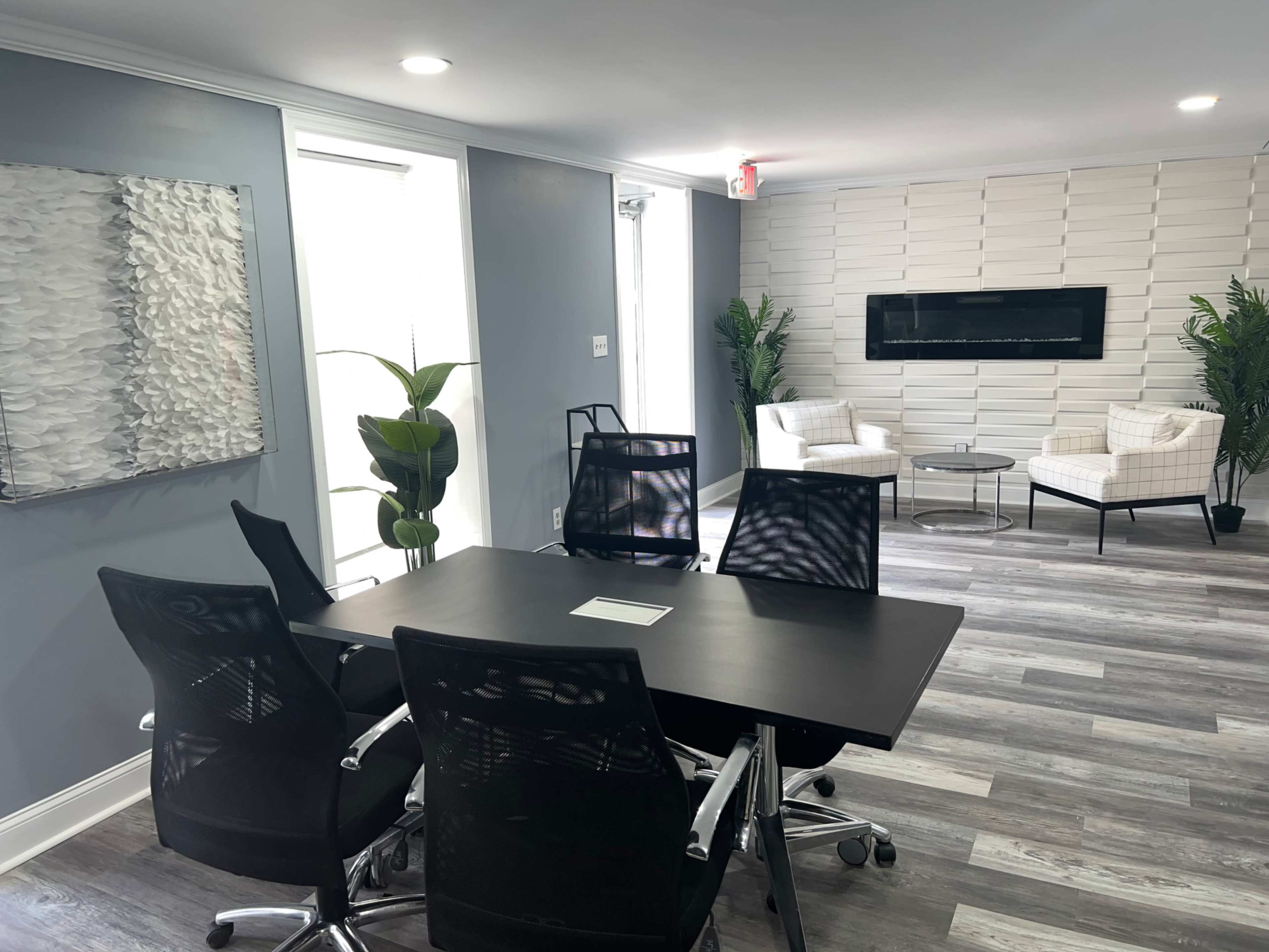 A modern office space features a meeting table surrounded by black chairs, with a comfortable seating area and a wall-mounted television in the background.