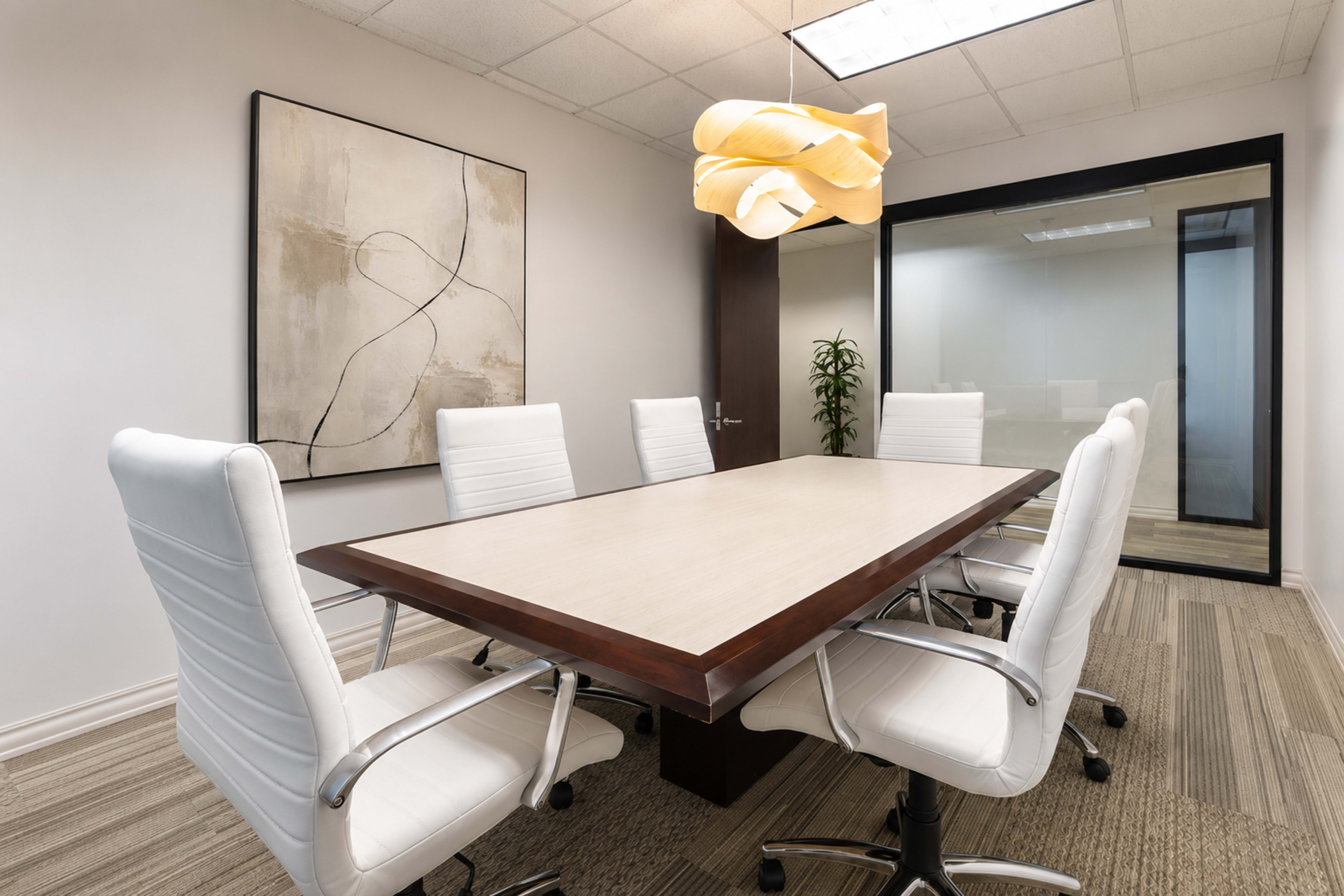 A modern conference room features a large rectangular table surrounded by six white leather chairs, with a light fixture overhead and a decorative wall piece in the background.