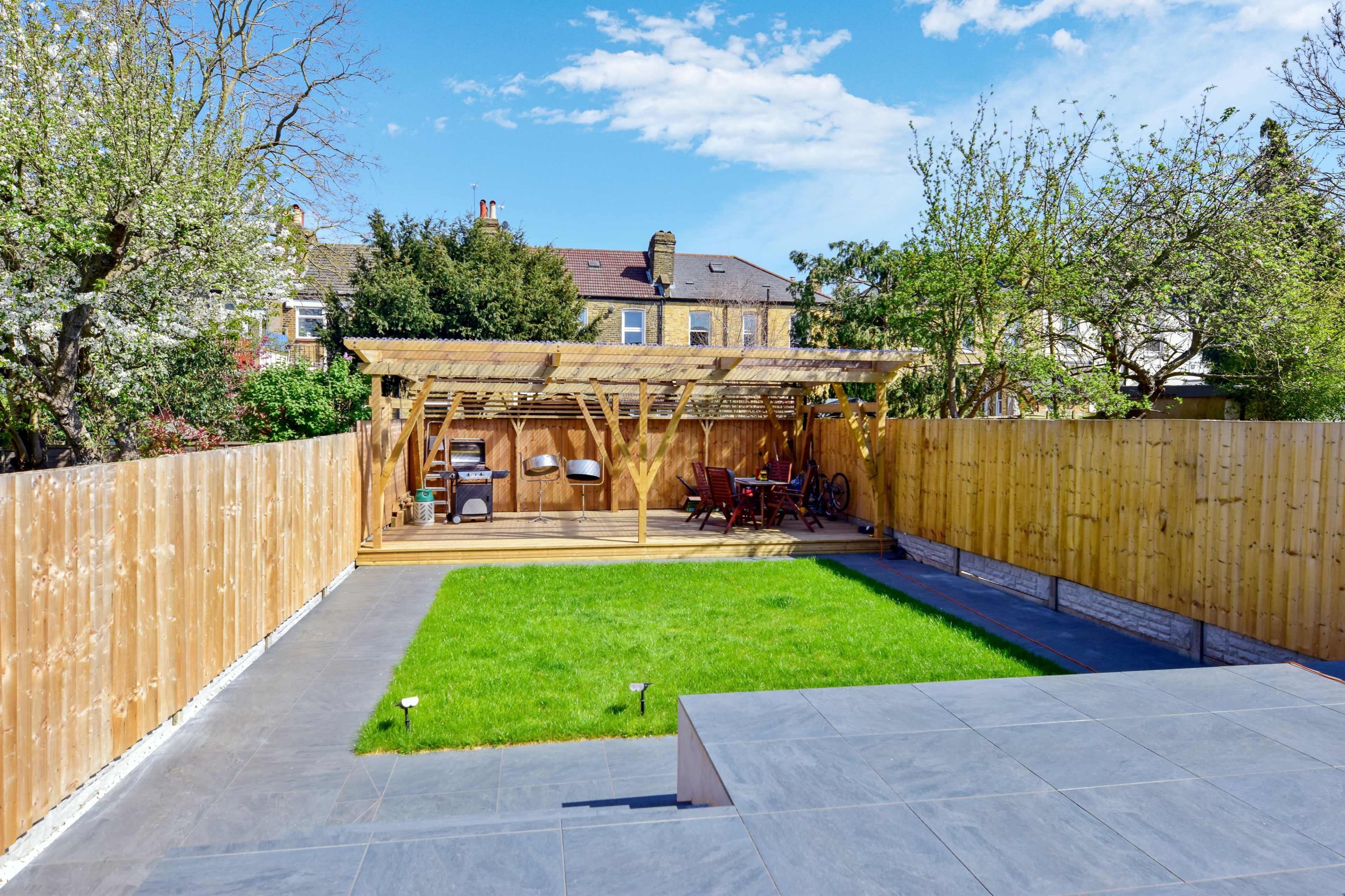 A well-maintained backyard featuring green grass, a wooden pergola with seating, and a fenced area surrounded by trees and a house in the background.