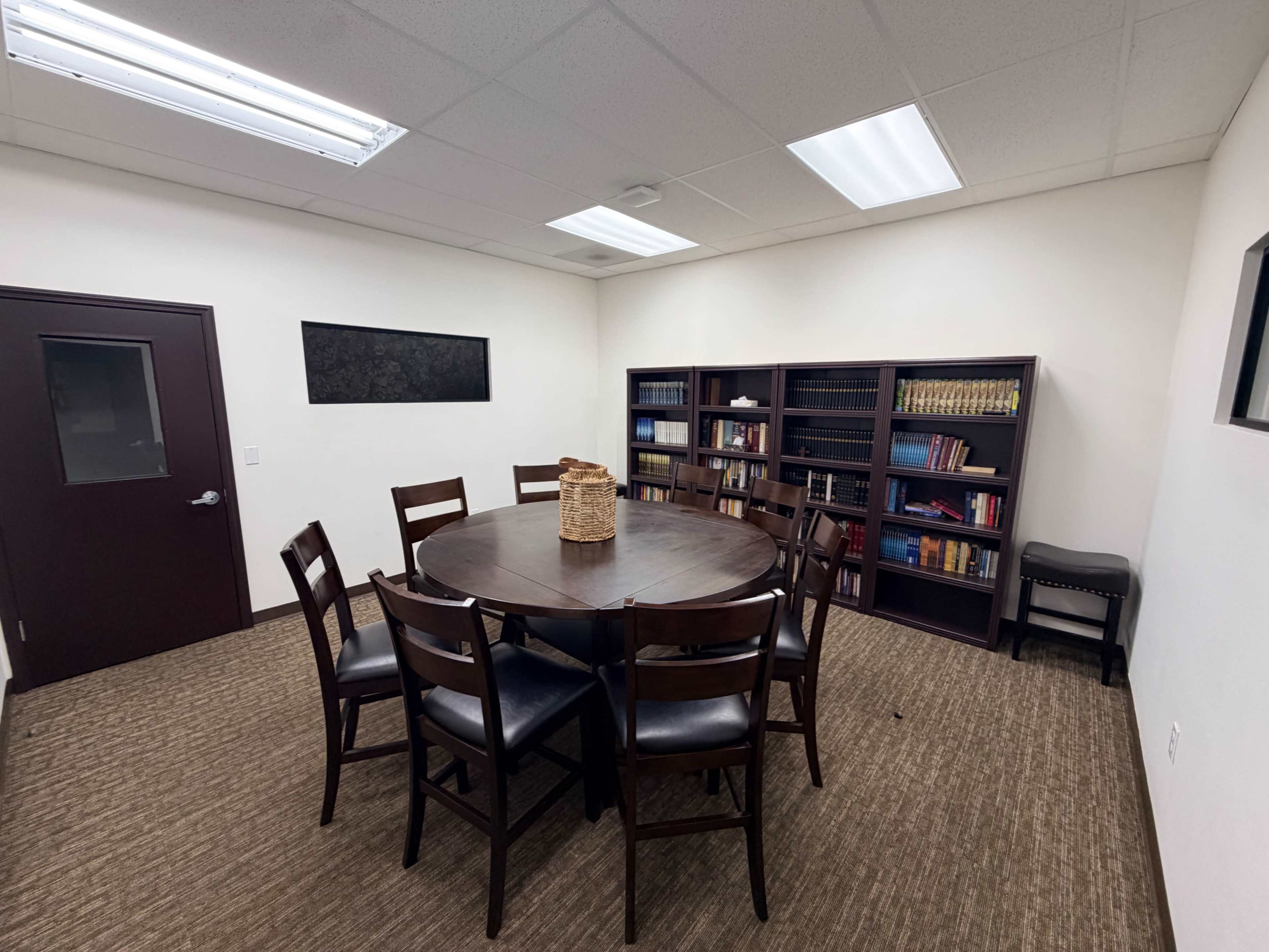 The image shows a small, well-lit meeting room containing a round wooden table surrounded by eight chairs, a bookshelf filled with books, and a door leading to another area.