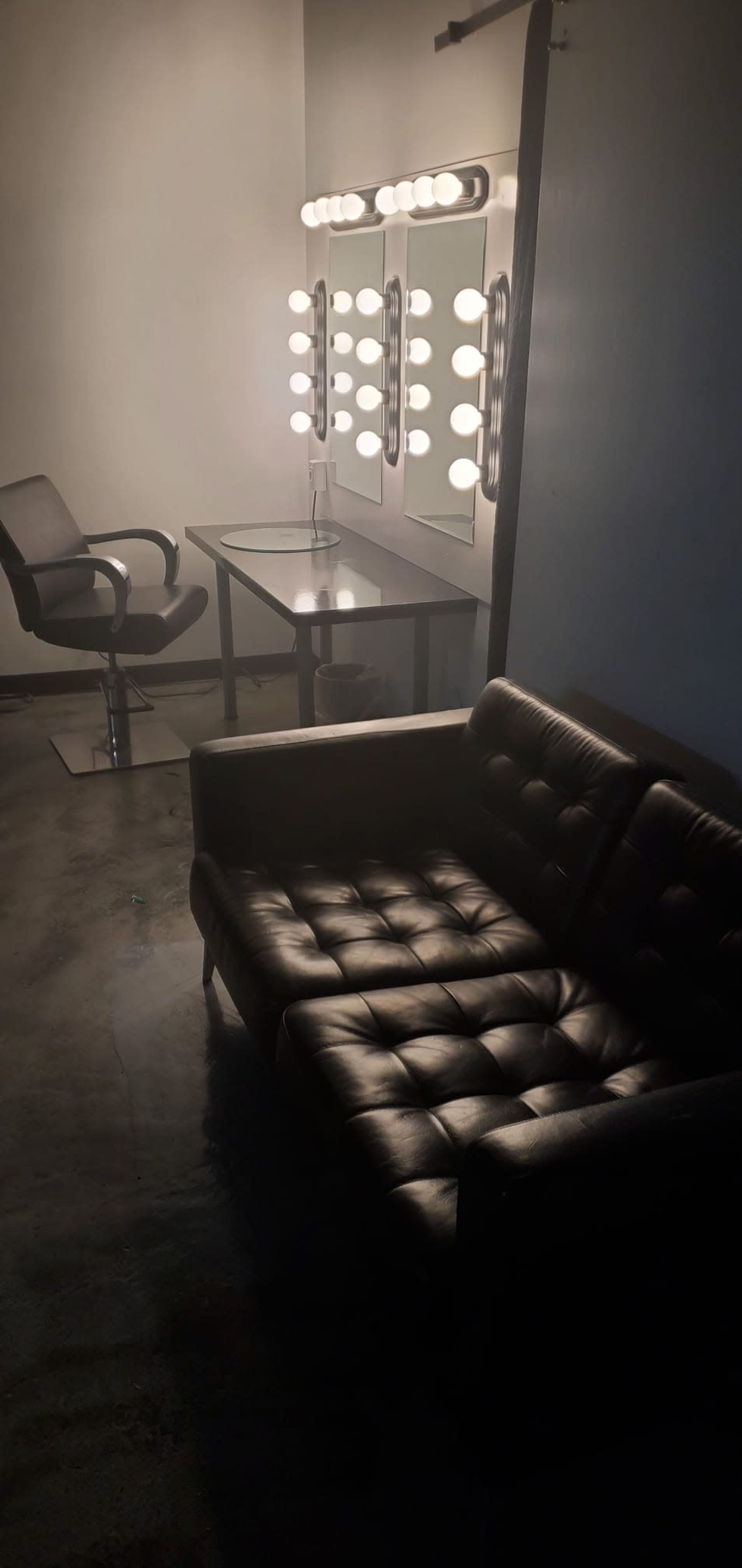 The image shows a dimly lit makeup studio featuring a black leather couch, a glass table, a salon chair, and a well-lit mirror with bulbs.