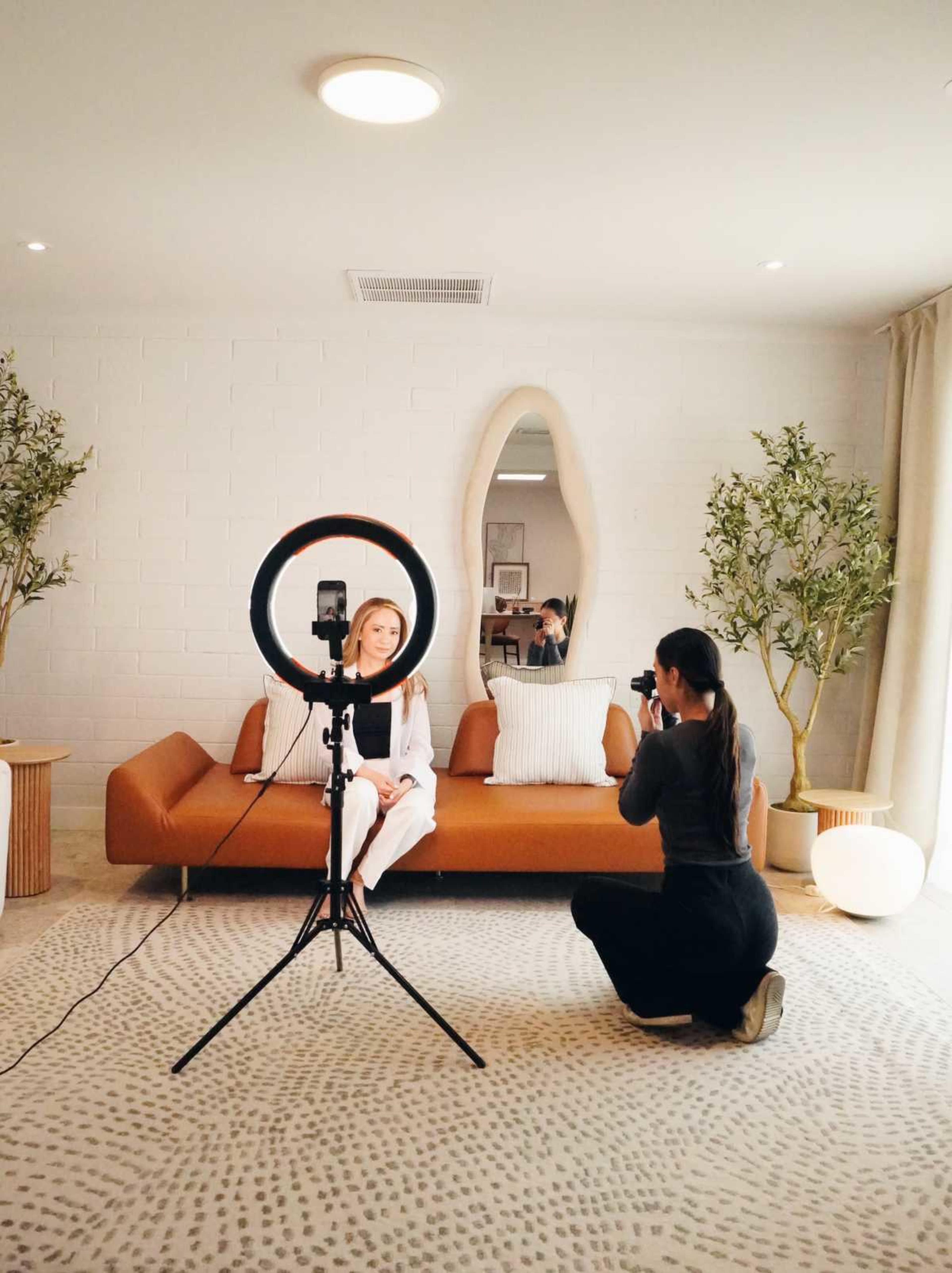 A woman is posing on a sofa in a light-filled room while another woman photographs her using a ring light.