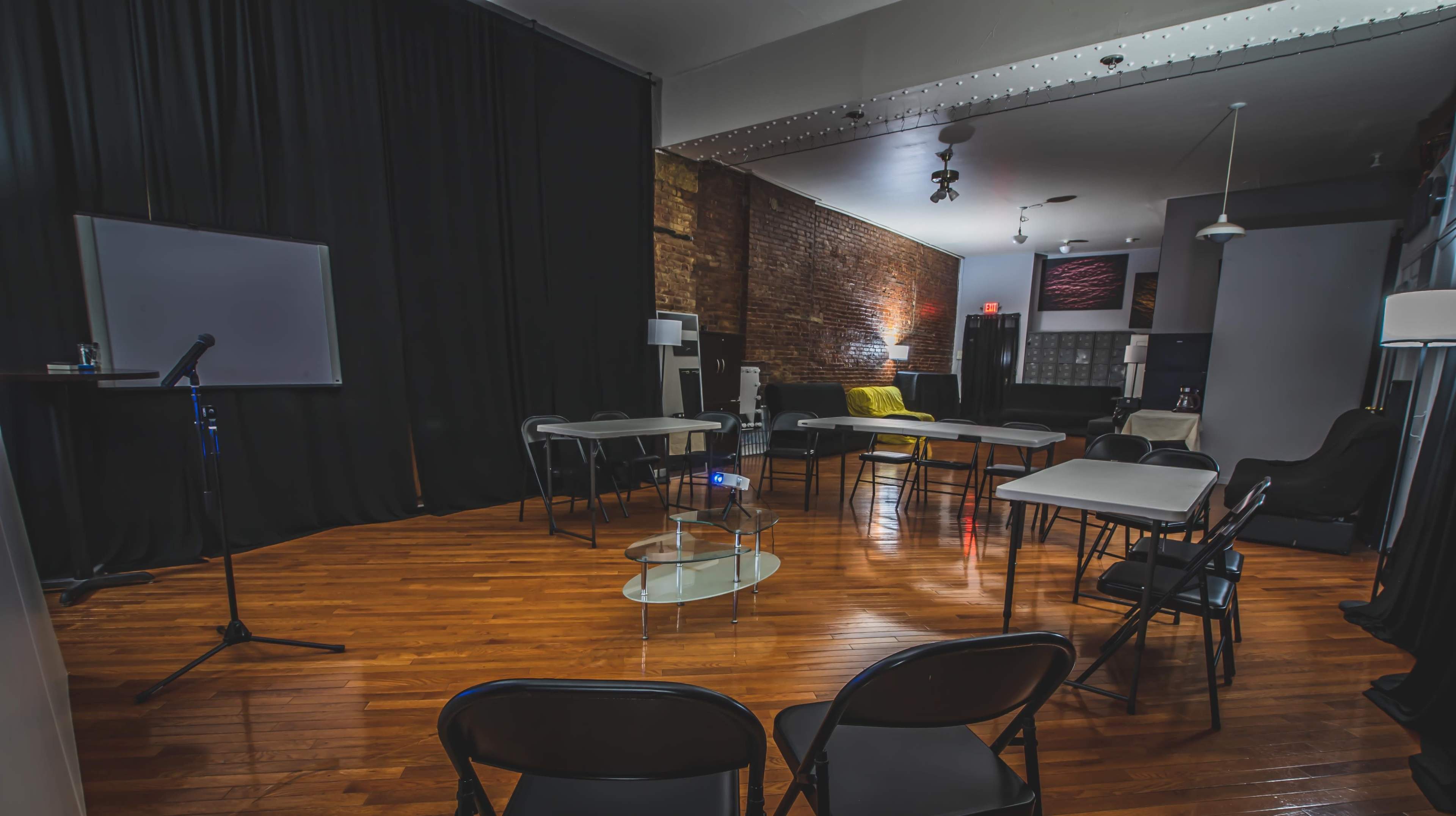 The image shows a room set up for an event, featuring several tables and chairs arranged in a circular format, with a projector and microphone visible at the front.