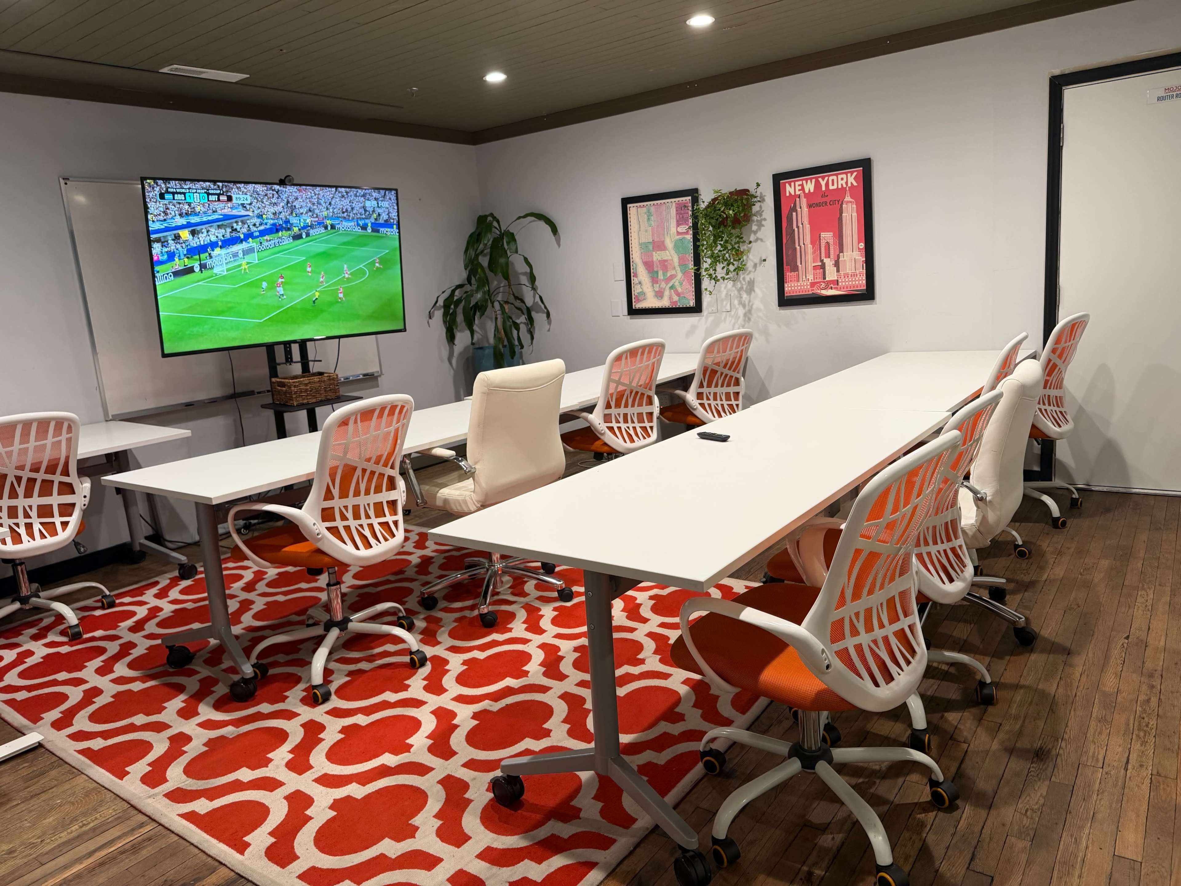 The image shows a modern conference room with a large screen displaying a soccer match, surrounded by white tables and chairs arranged in a semi-circle on a red patterned rug.