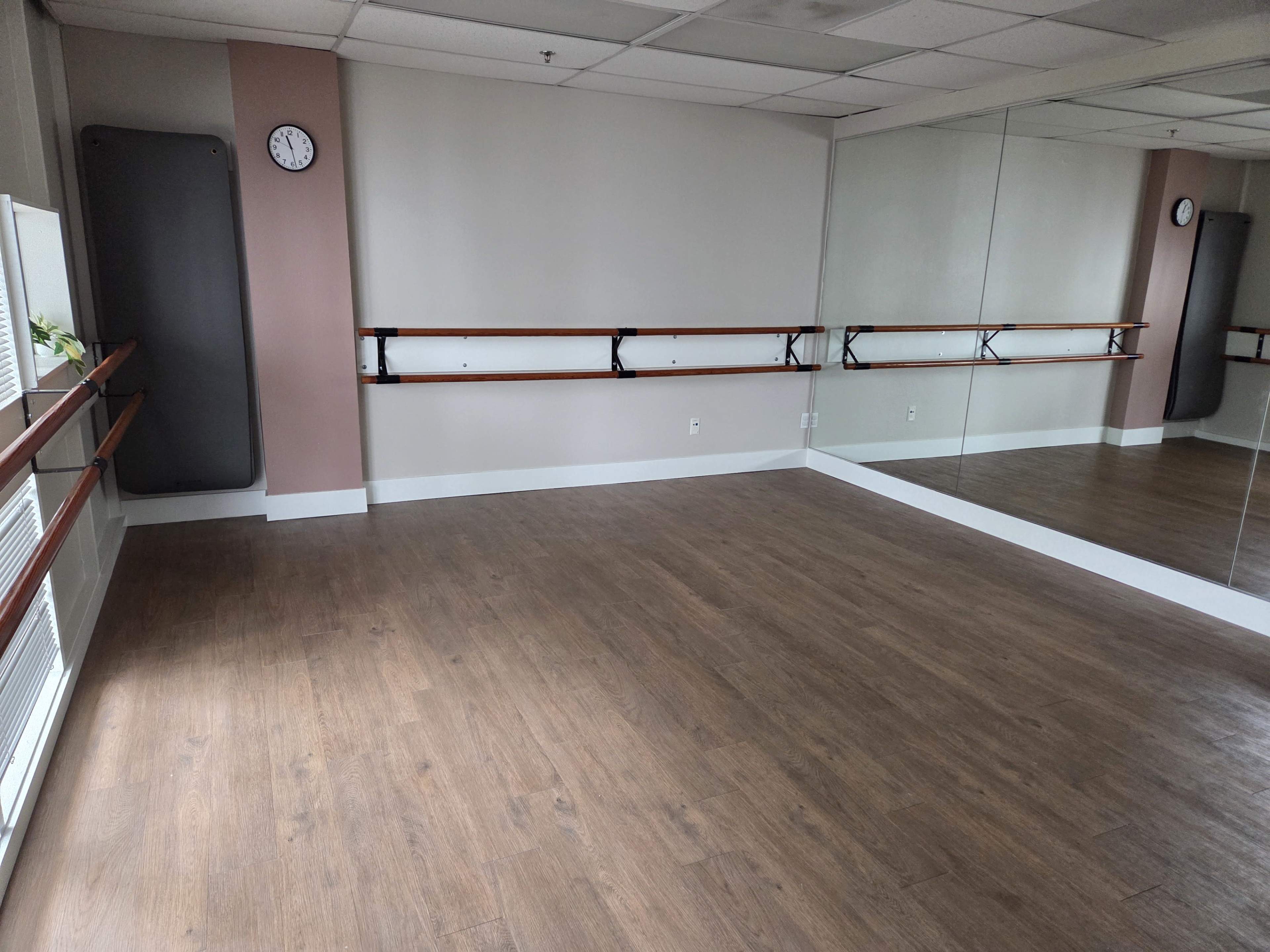 The image shows an empty dance studio with wooden flooring, mirrors on one wall, and a wall-mounted clock.