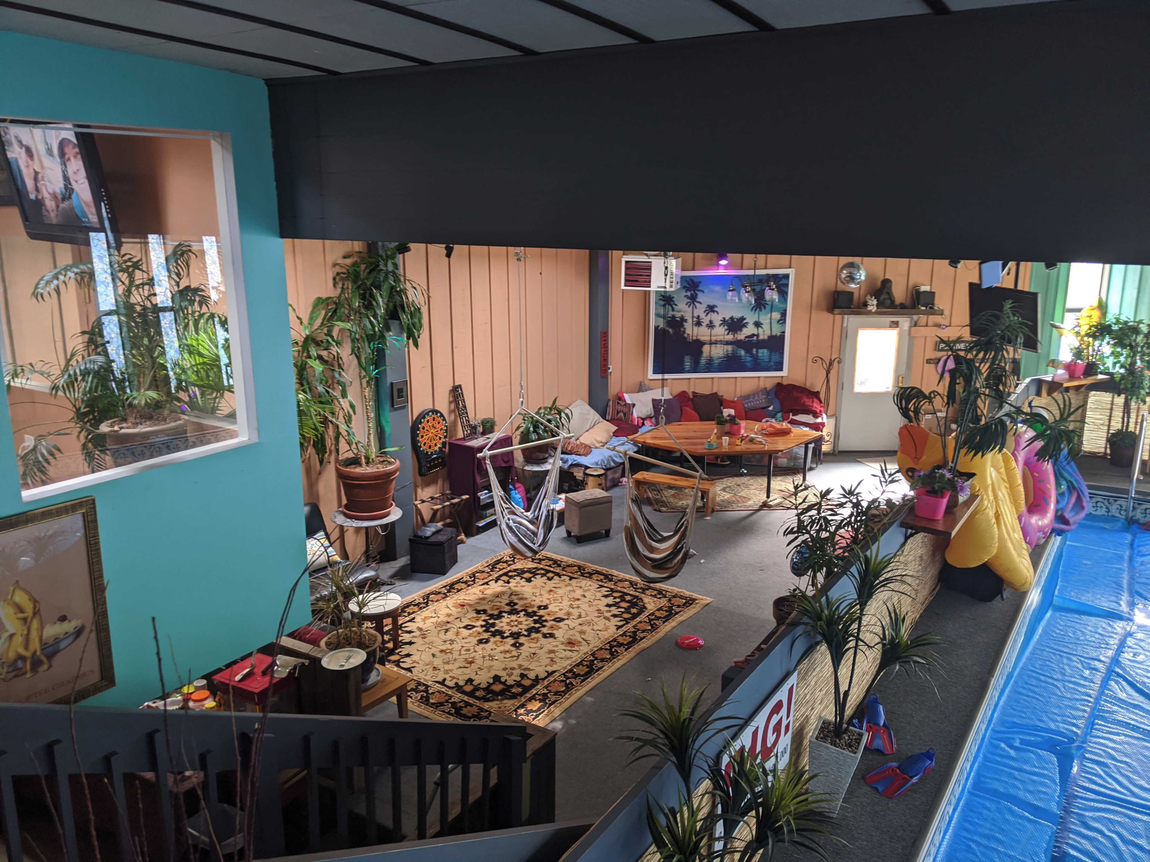 A cozy indoor space with a large round table, a patterned rug, various plants, and a poolside area visible on one side.