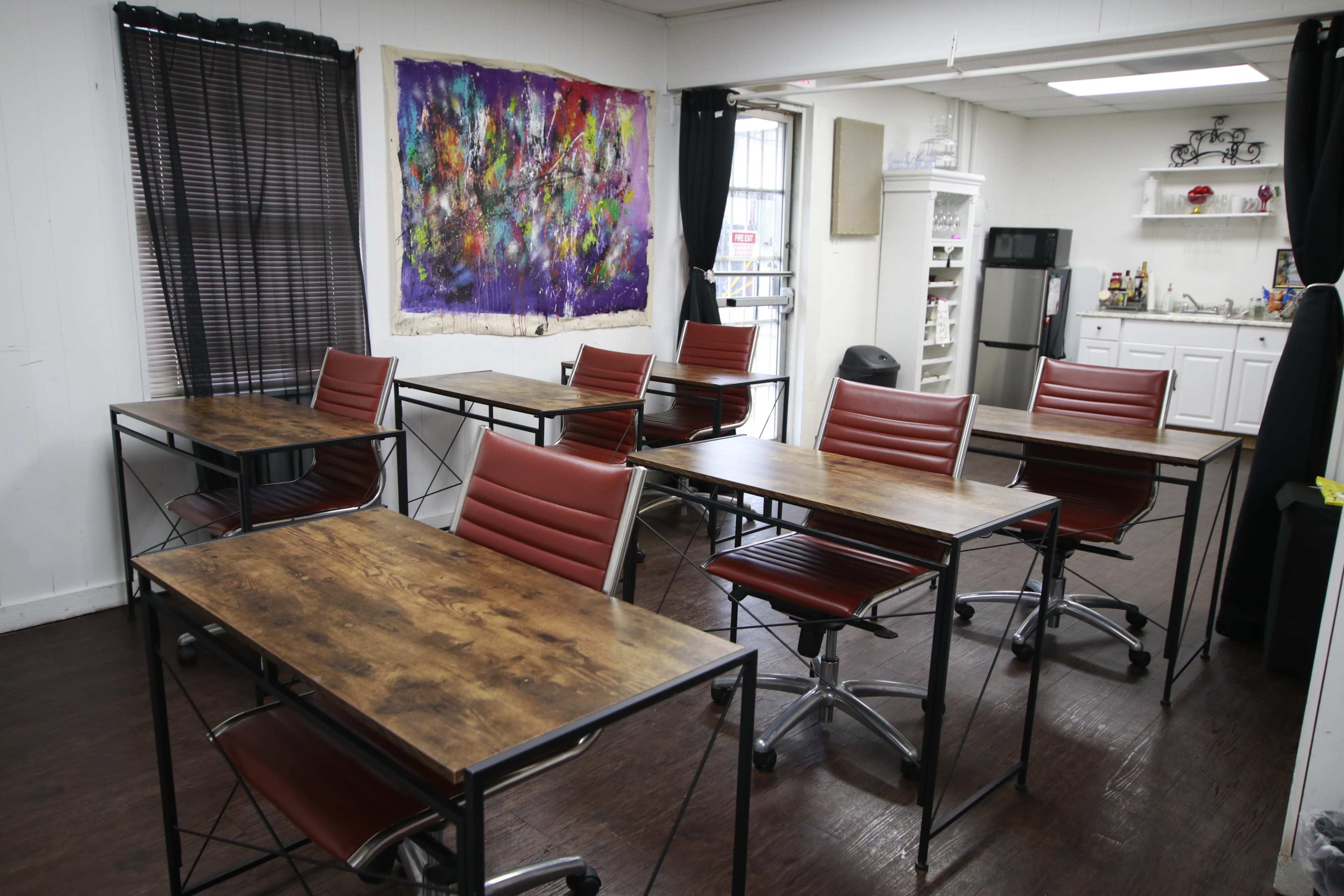 A classroom setup with multiple wooden tables and red chairs, flanked by black curtains and a colorful artwork on the wall.