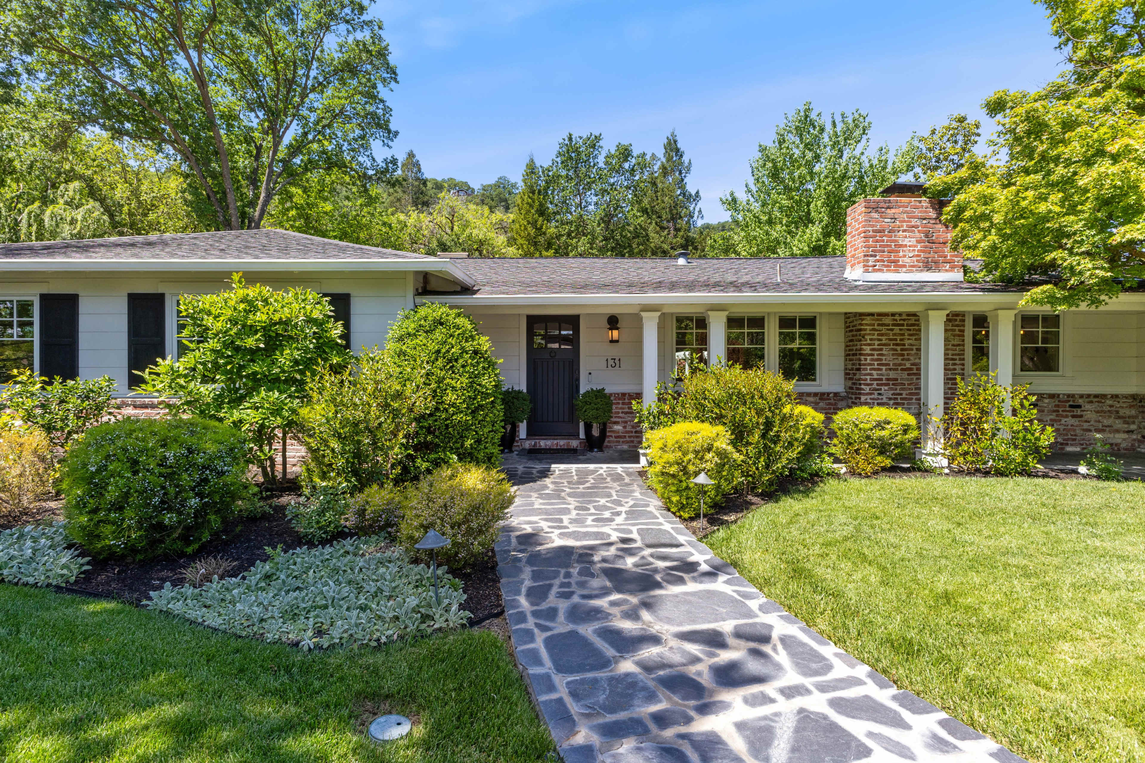 A single-story house with a stone walkway and well-maintained landscaping is surrounded by trees and shrubs.