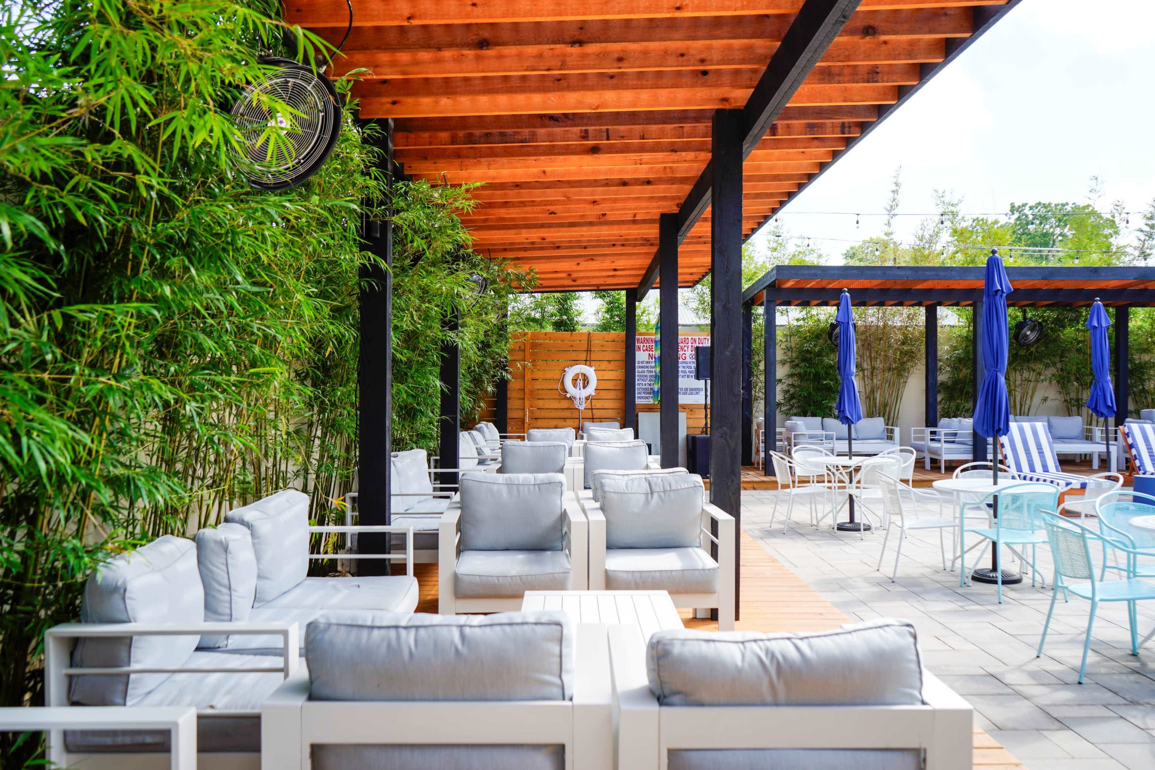 The image shows an outdoor seating area with gray cushioned furniture under a wooden pergola, surrounded by bamboo and tables with umbrellas.