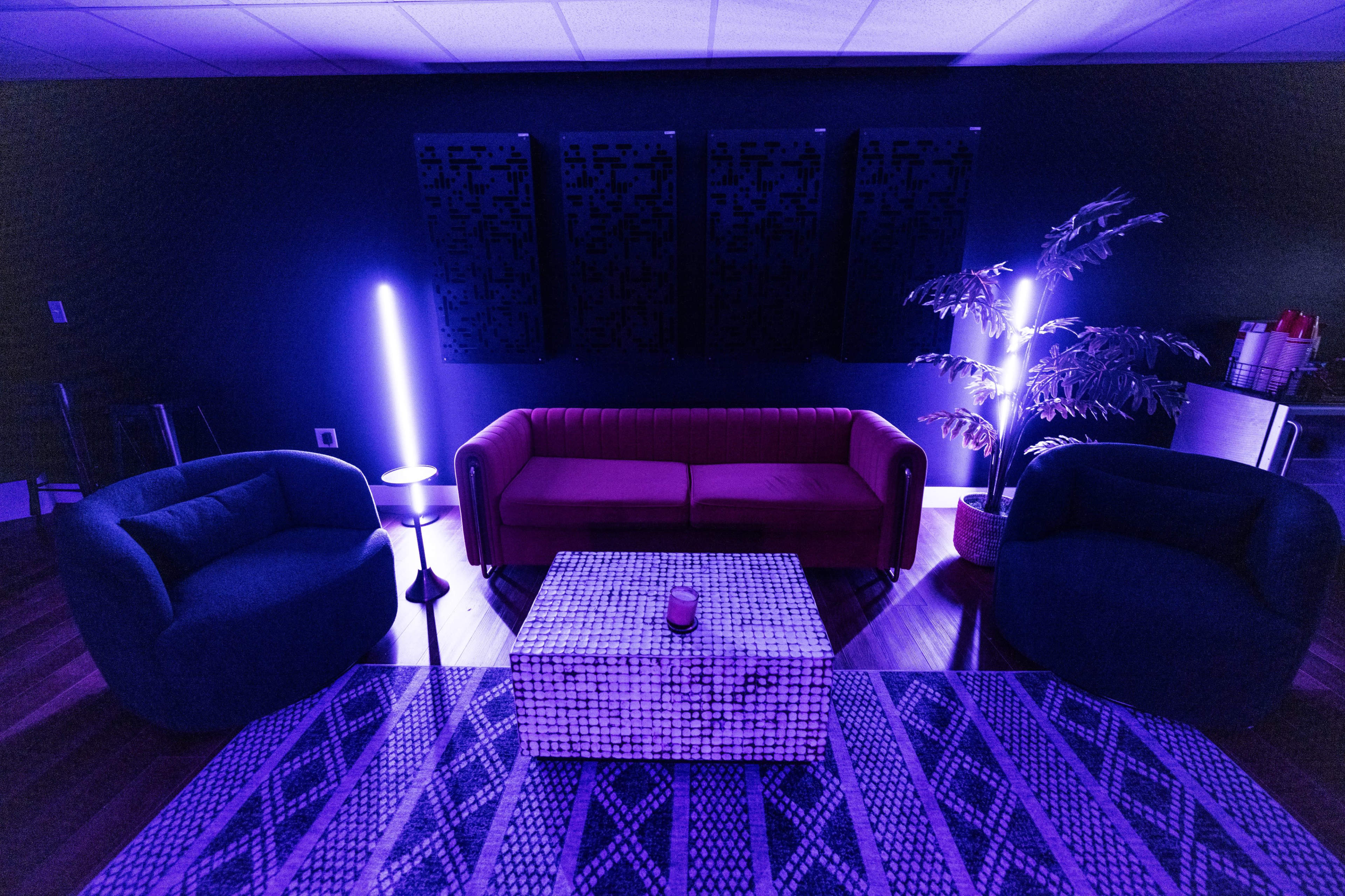 A modern lounge area features a red sofa, two blue armchairs, a patterned coffee table, and illuminated by purple lights.