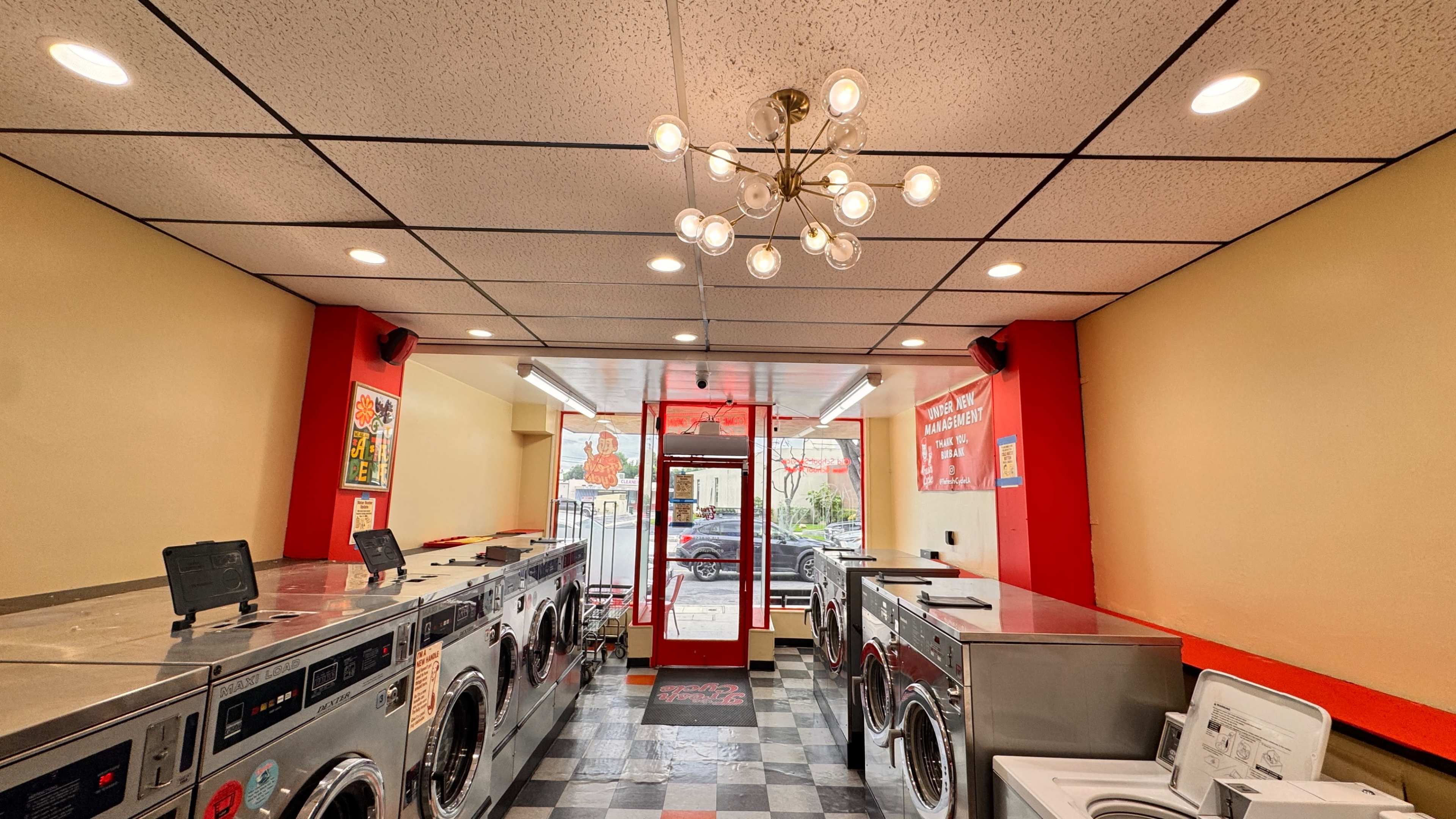 The interior of a laundromat features rows of washer and dryer machines, a seating area, and large windows displaying a view of the outside.