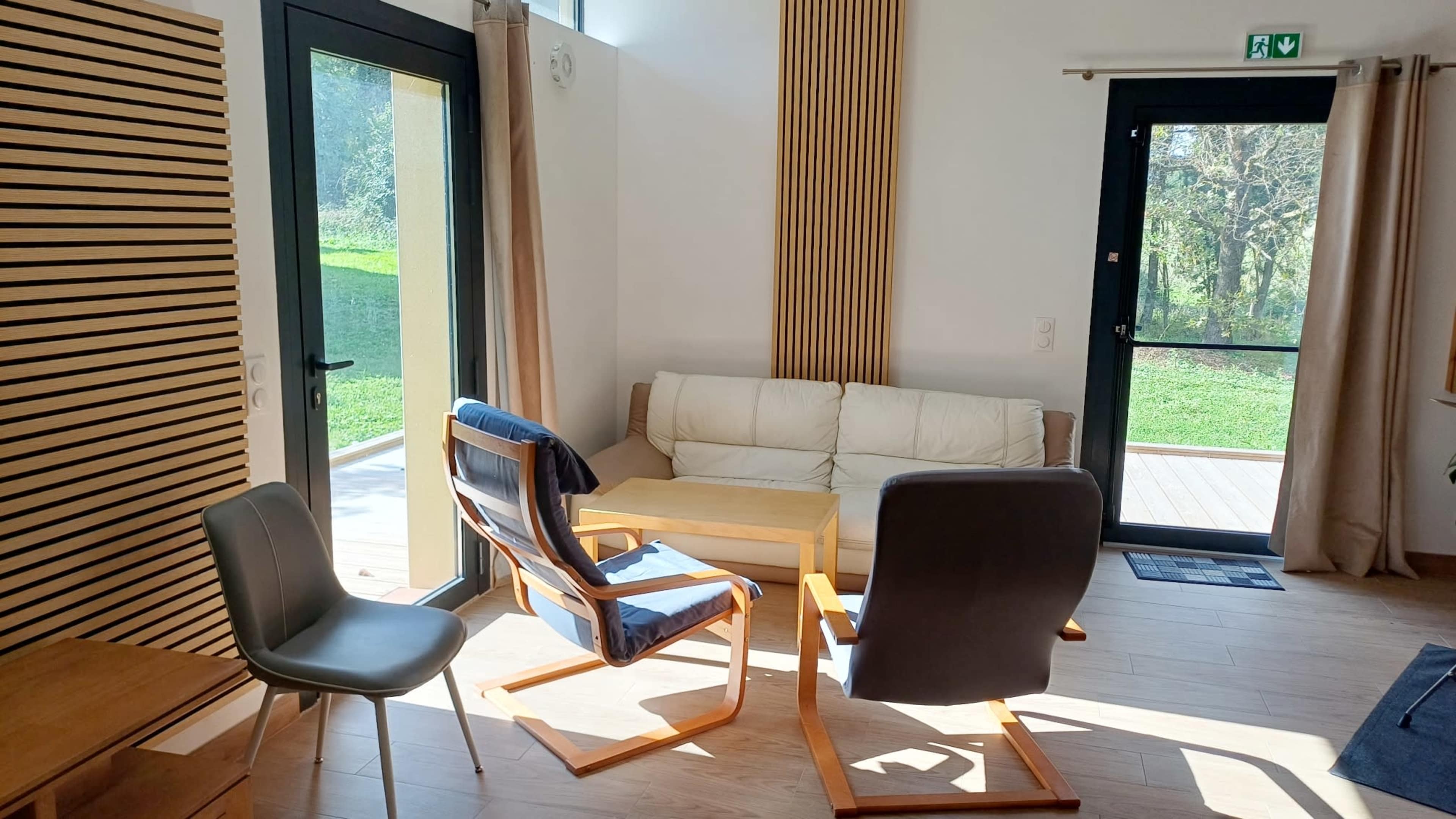 The image shows a bright interior room with a white sofa, a wooden coffee table, and two chairs positioned around it, with large windows opening to a green outdoor view.