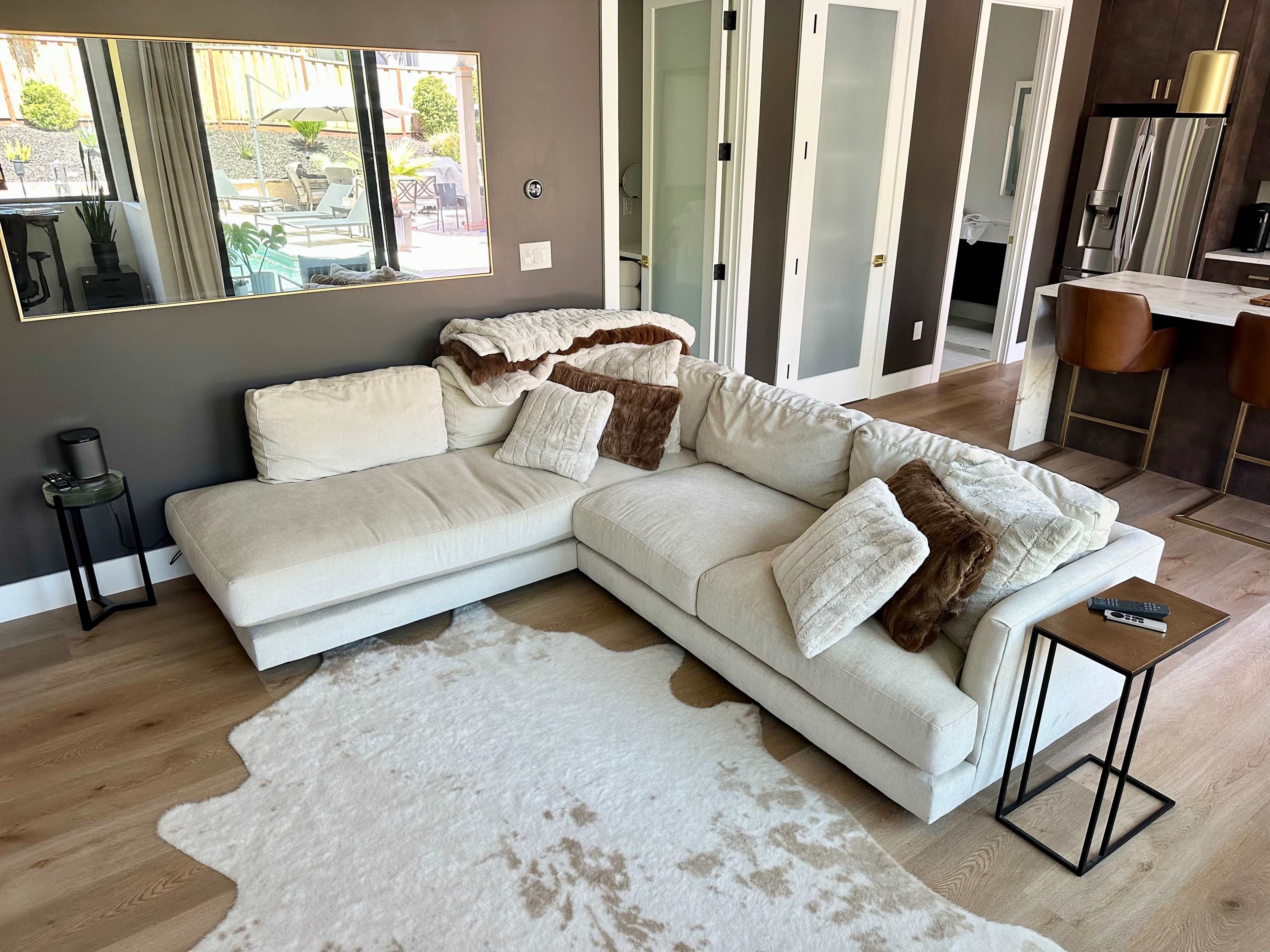 A modern living room with a spacious beige sectional sofa, decorative pillows, a cowhide rug, and a mirrored wall, leading to a bright outdoor area visible through glass doors.