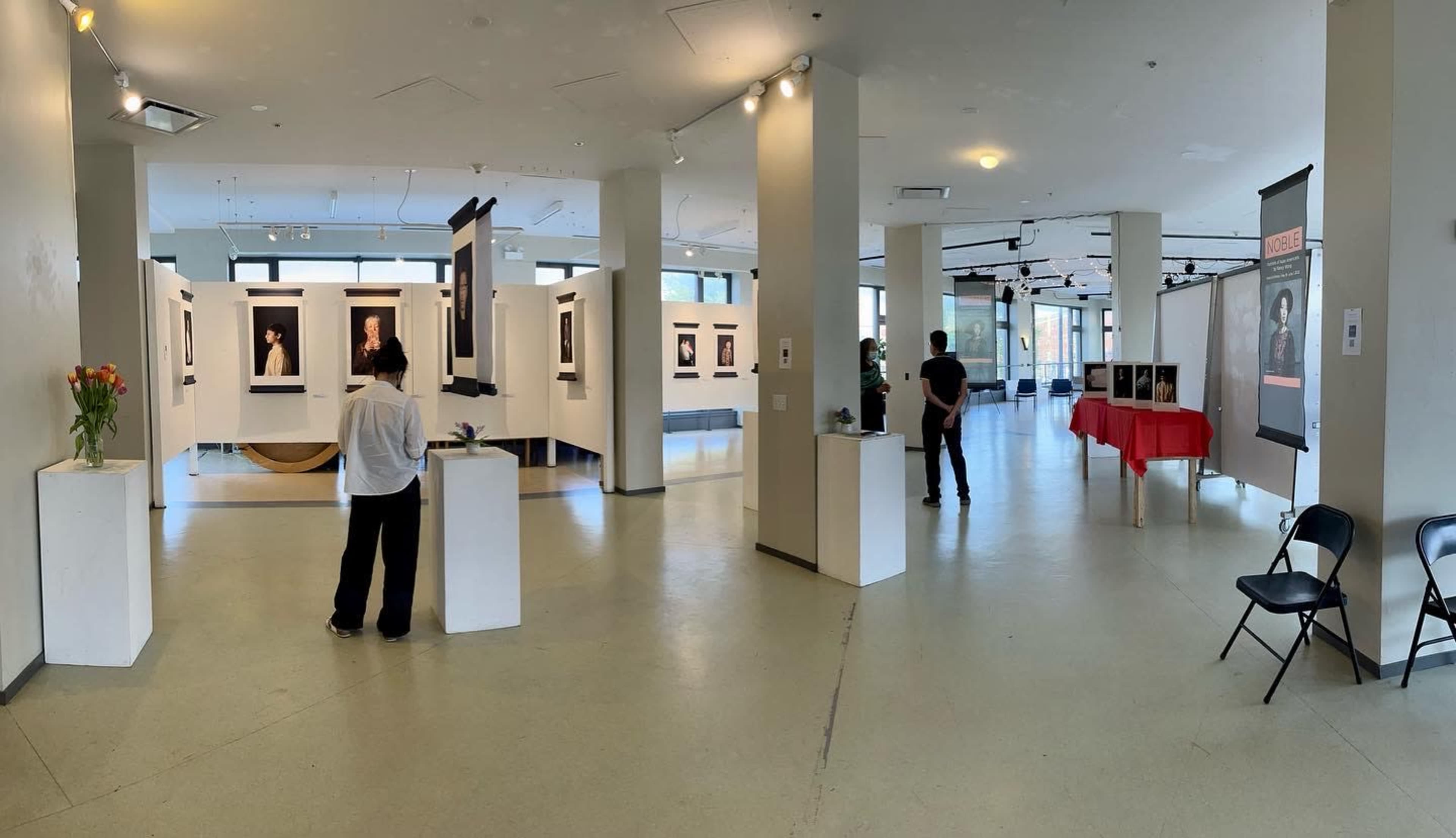 The photo shows an art gallery space with multiple framed portraits displayed on the walls and visitors observing the artwork.