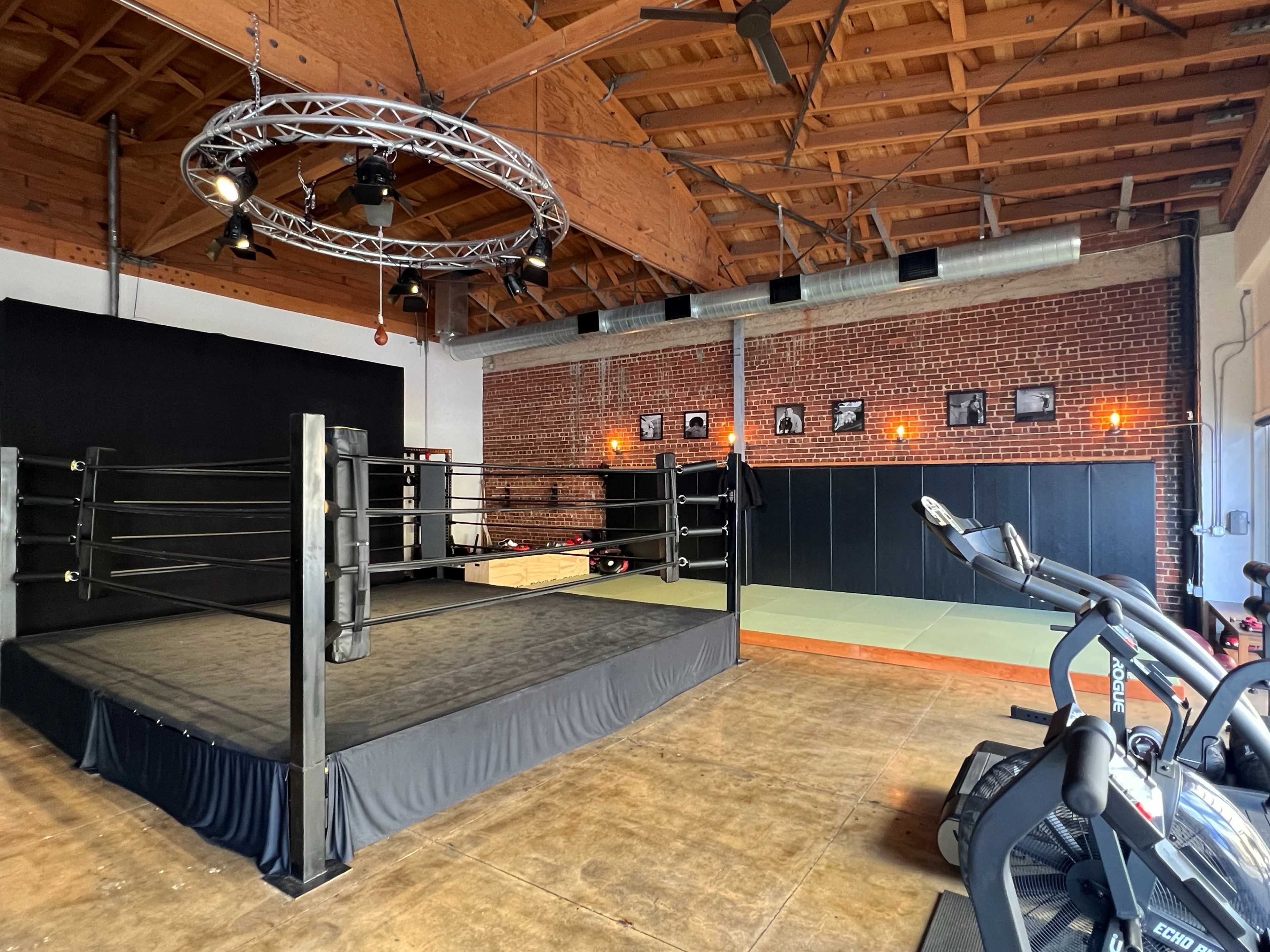 An indoor training facility featuring a boxing ring, exercise equipment, and a brick wall with framed photos.