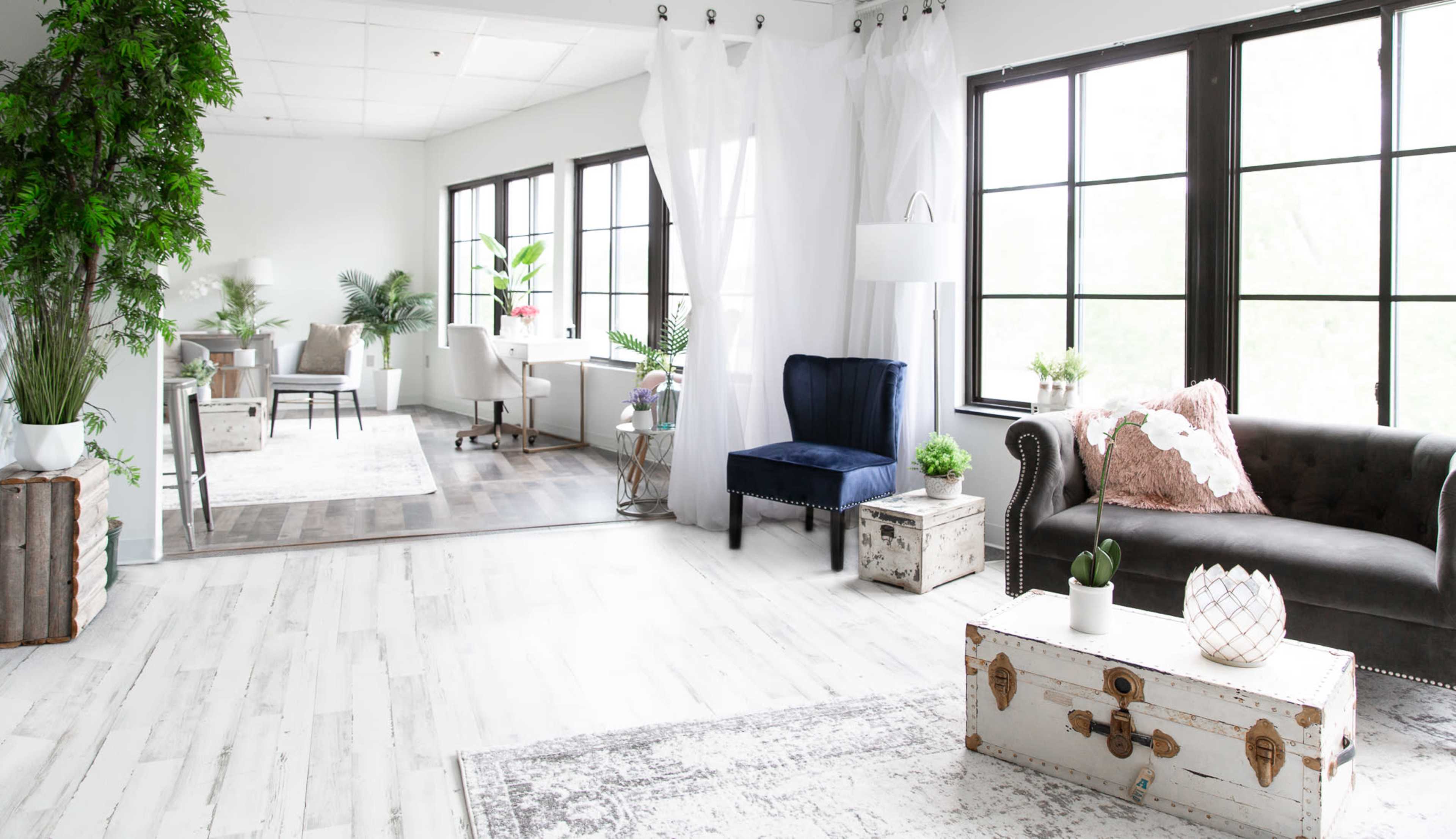 A bright, modern interior space with white walls, large windows, and a mix of furniture including a velvet chair, a sofa, and various plants.