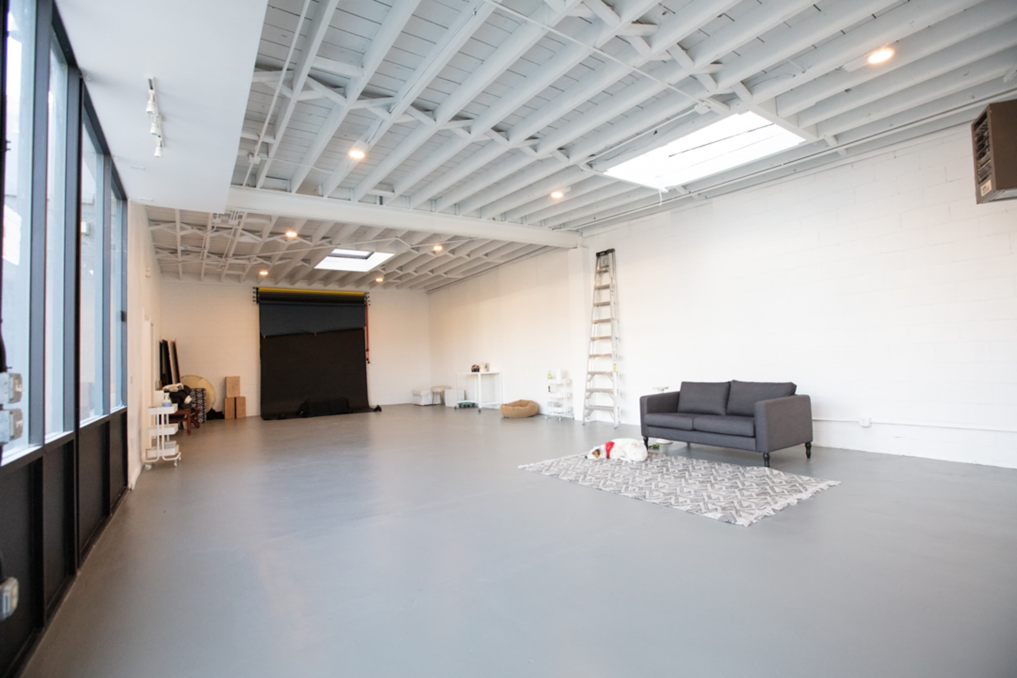 A spacious, empty studio with a gray floor, white walls, a sofa, and a large window allowing natural light to enter.