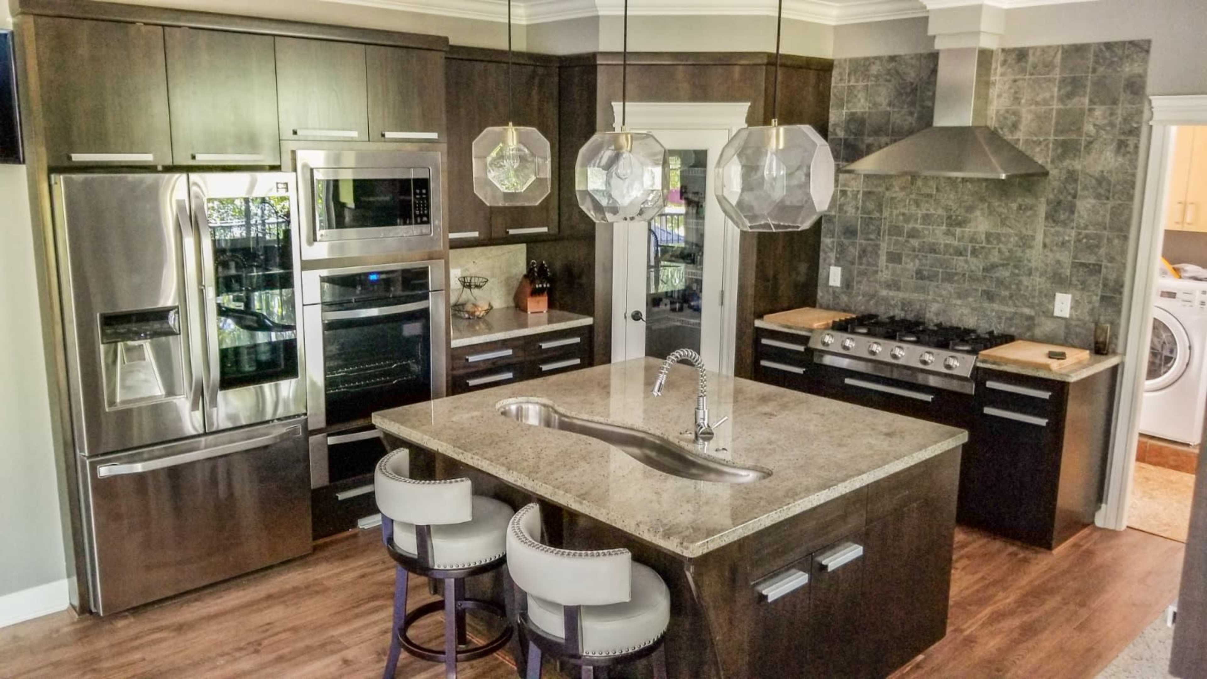 A modern kitchen featuring dark wood cabinets, stainless steel appliances, a large granite island with seating, and pendant lighting.
