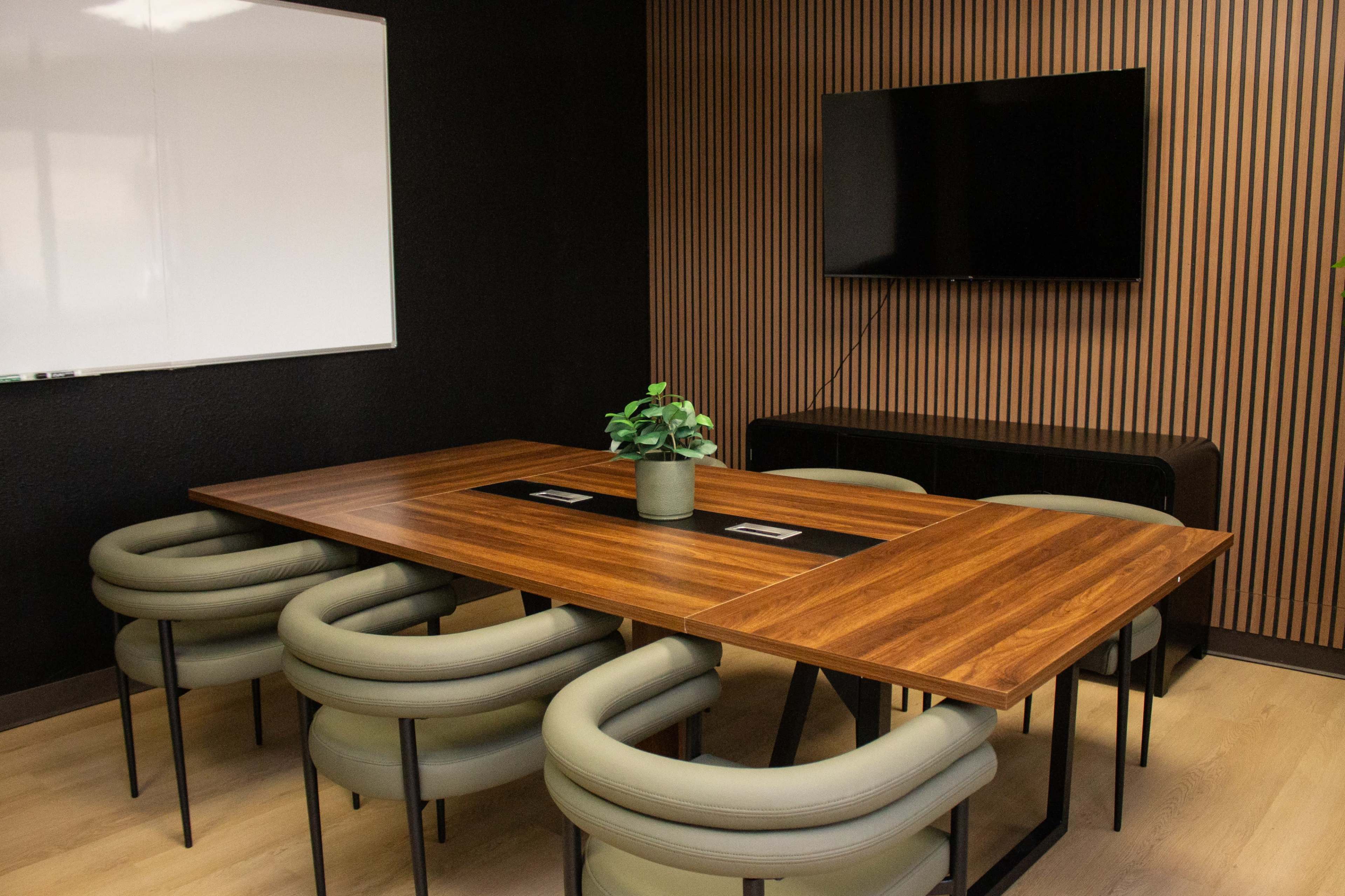 The image shows a modern conference room featuring a wooden table surrounded by six circular chairs, a flat-screen TV on the wall, and a potted plant in the center.