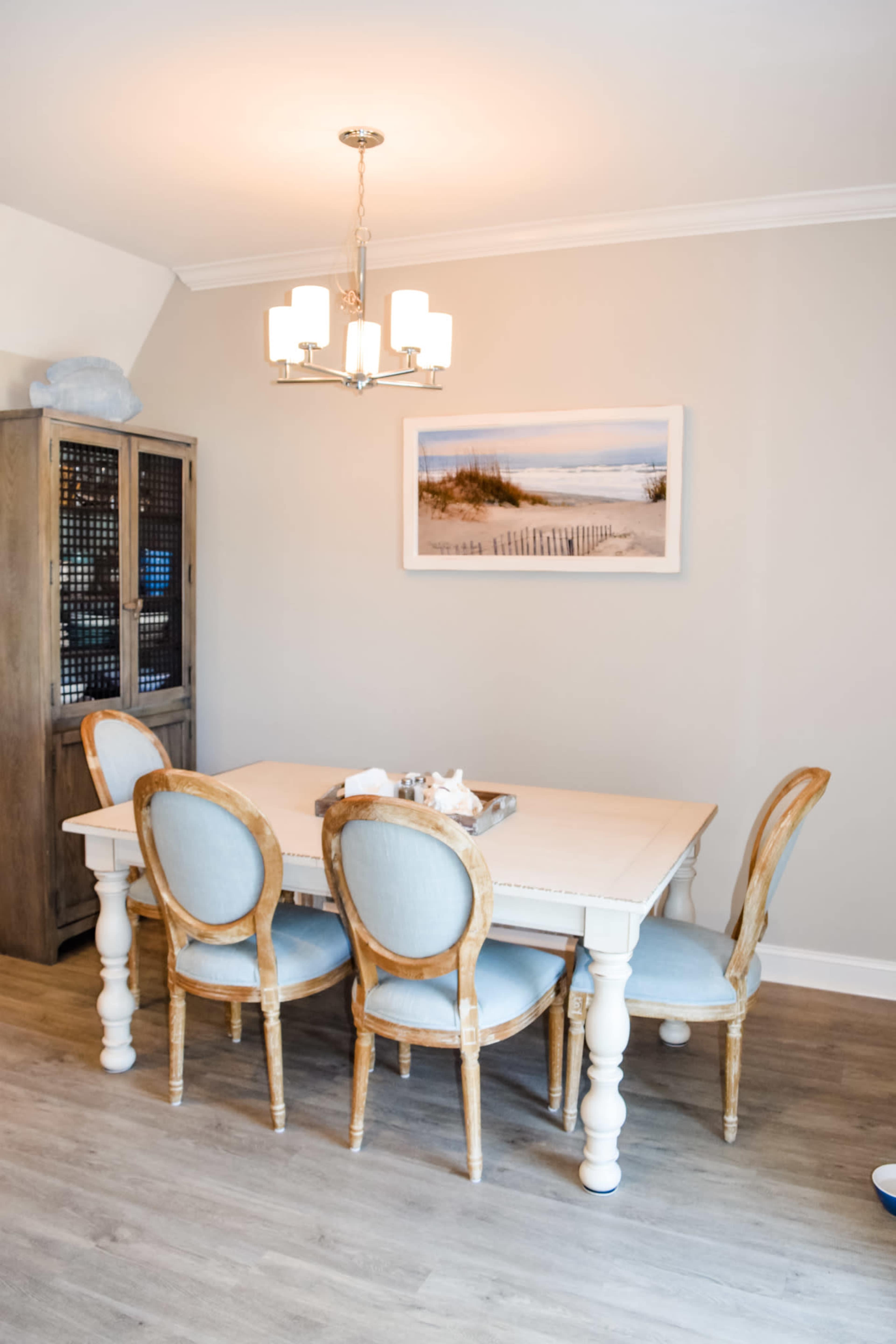 A light-colored dining room features a wooden table surrounded by four upholstered chairs, with a photograph of a beach scene hanging on the wall above.