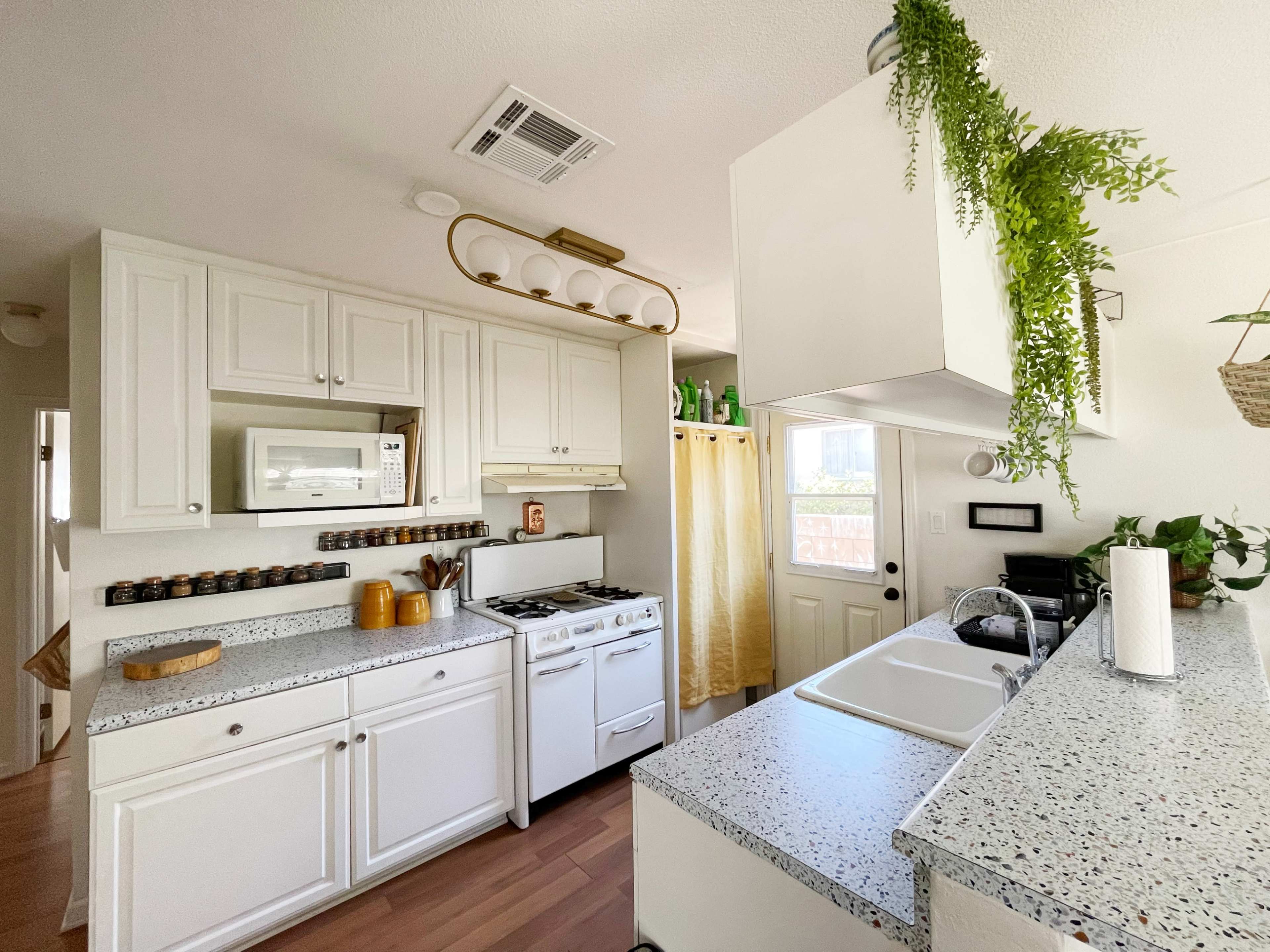 A bright kitchen features white cabinets, a stove, a microwave, and a sink, with a green plant hanging from the ceiling and wooden accents.