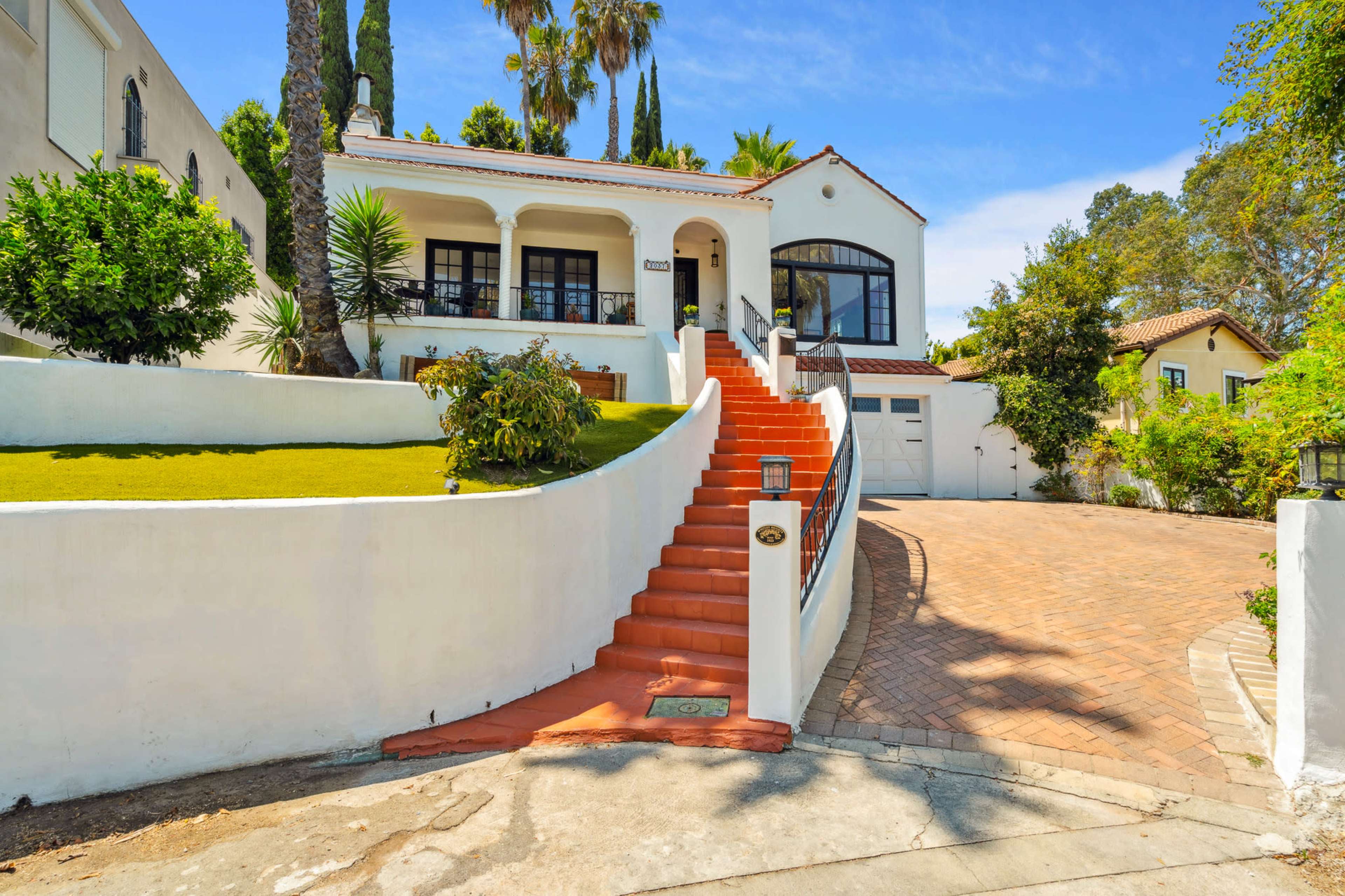 A white house with a red-tiled roof sits on a landscaped lot, featuring a curved orange staircase leading to the entrance and a driveway made of pavers.