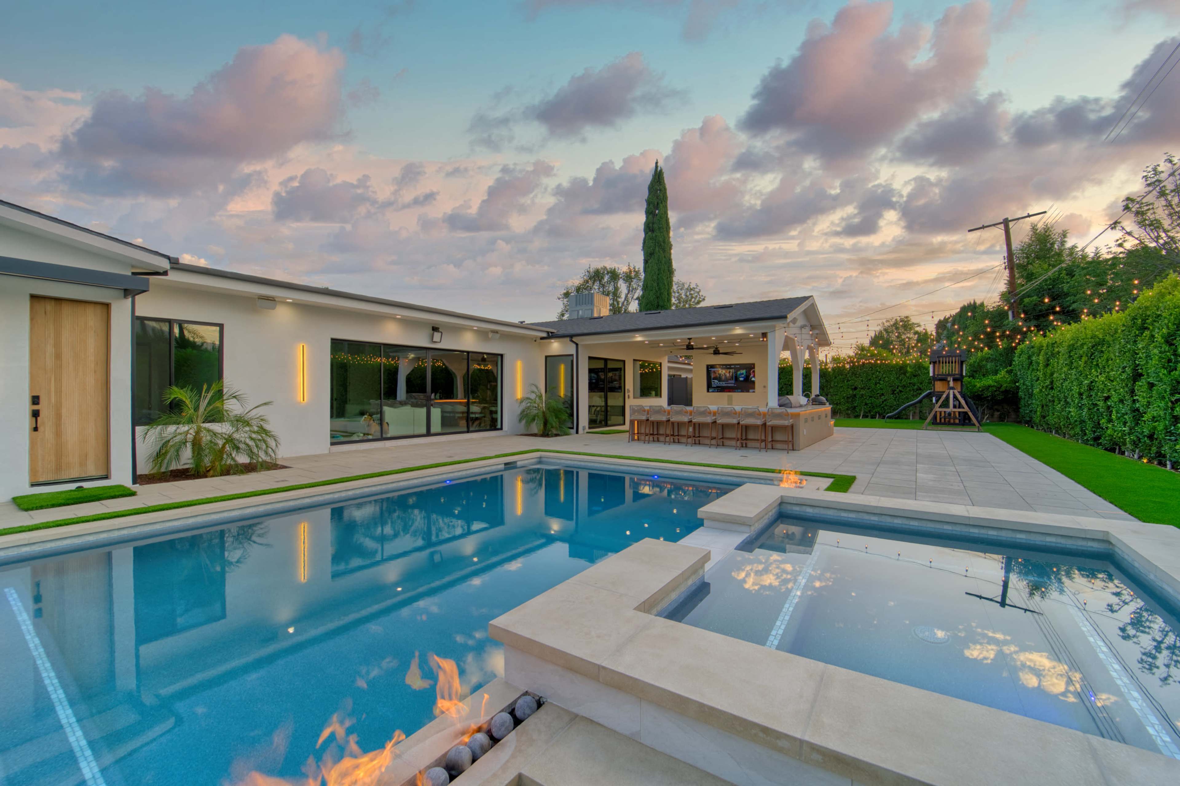 The image shows a modern backyard featuring a swimming pool with fire features, a patio area, and a well-manicured landscape under a cloudy sky.