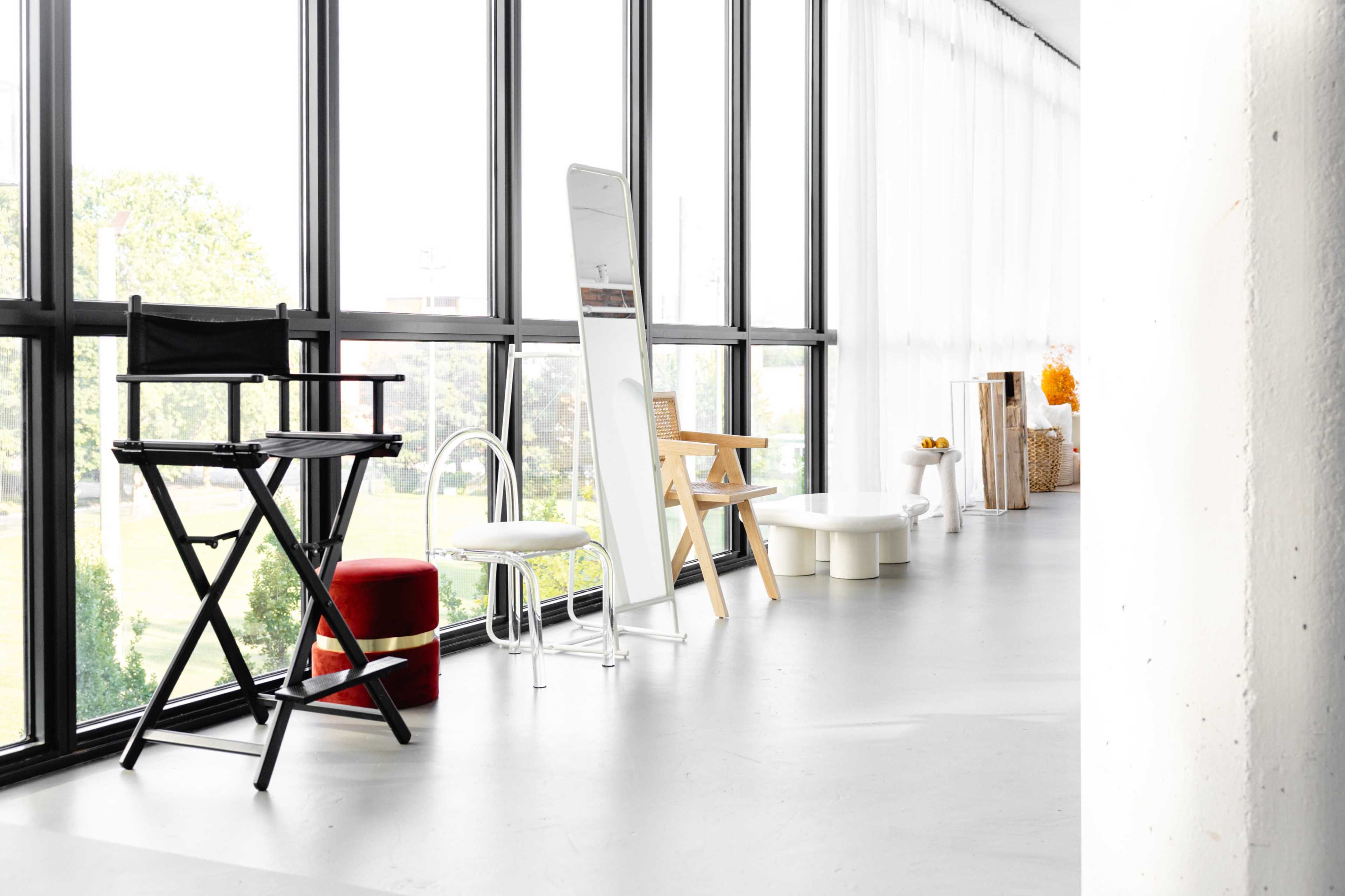The image shows a modern, spacious interior with large windows, featuring various pieces of furniture including a director's chair, a white mirror, and several other seating options arranged along a light-colored floor.