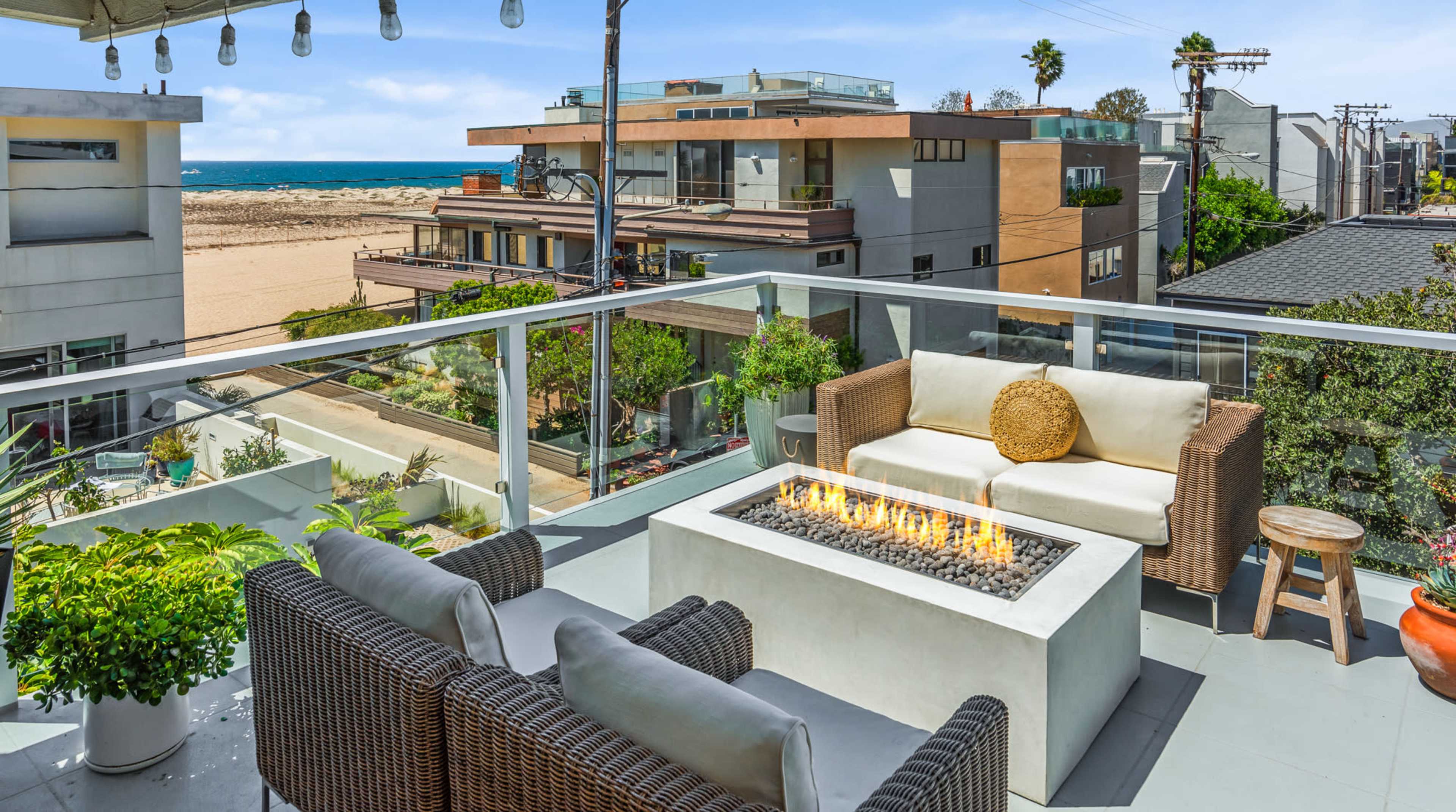 A balcony features a fire pit surrounded by outdoor seating, overlooking a sandy beach and modern buildings.
