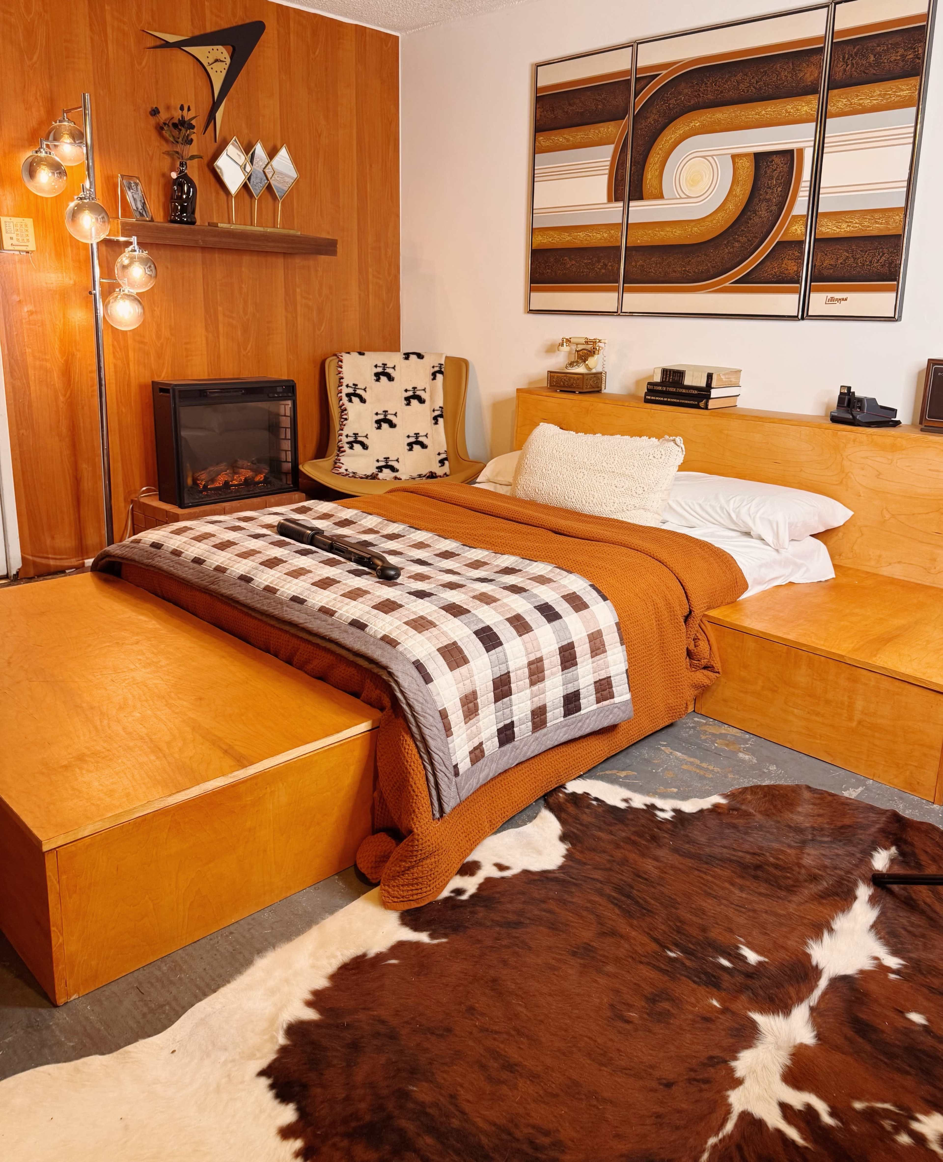 The image shows a cozy bedroom featuring wooden furniture, a bed with an orange blanket, a patterned throw, and a cowhide rug on the floor.