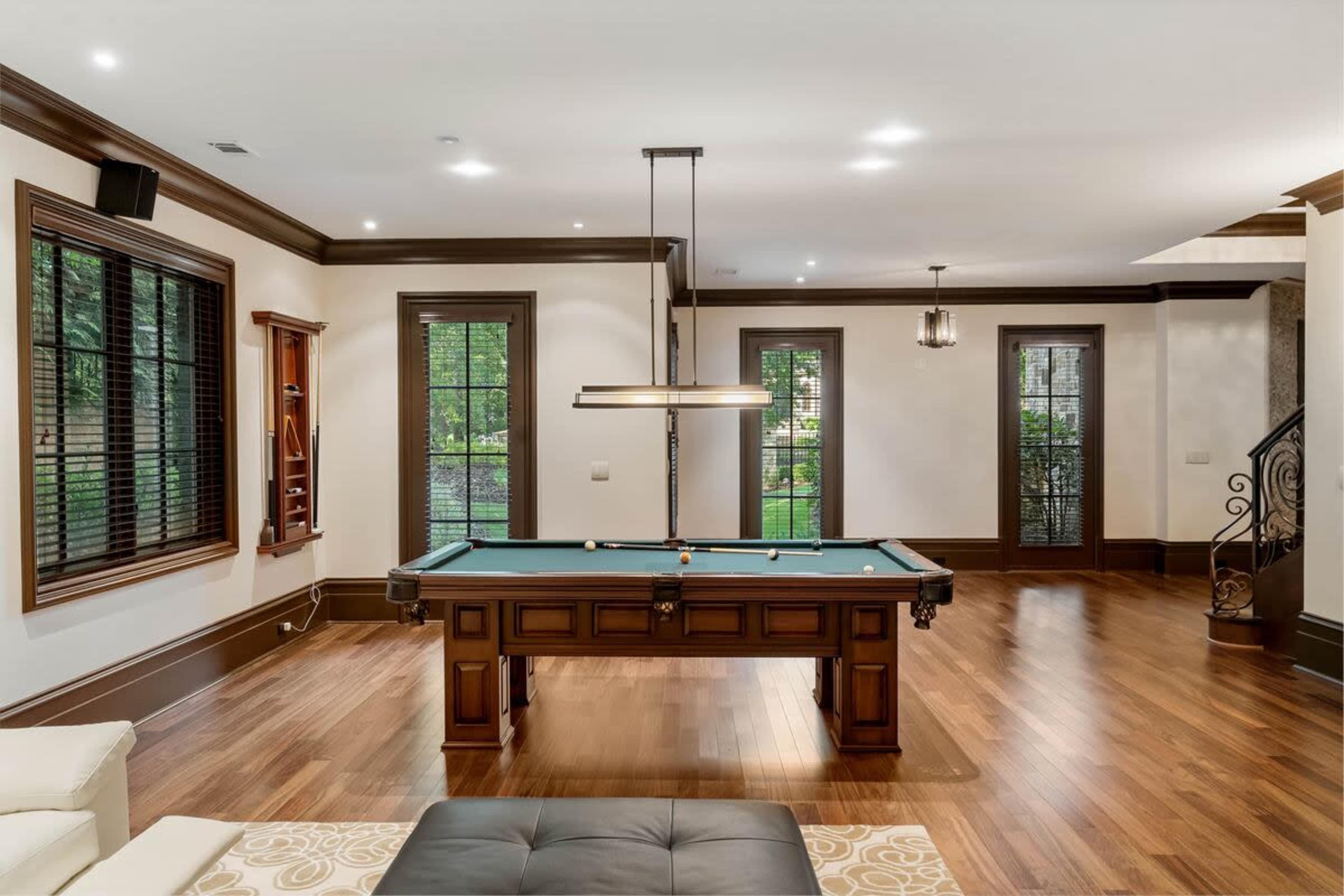 The image shows a spacious game room with a billiards table, hardwood flooring, and large windows allowing natural light to enter.