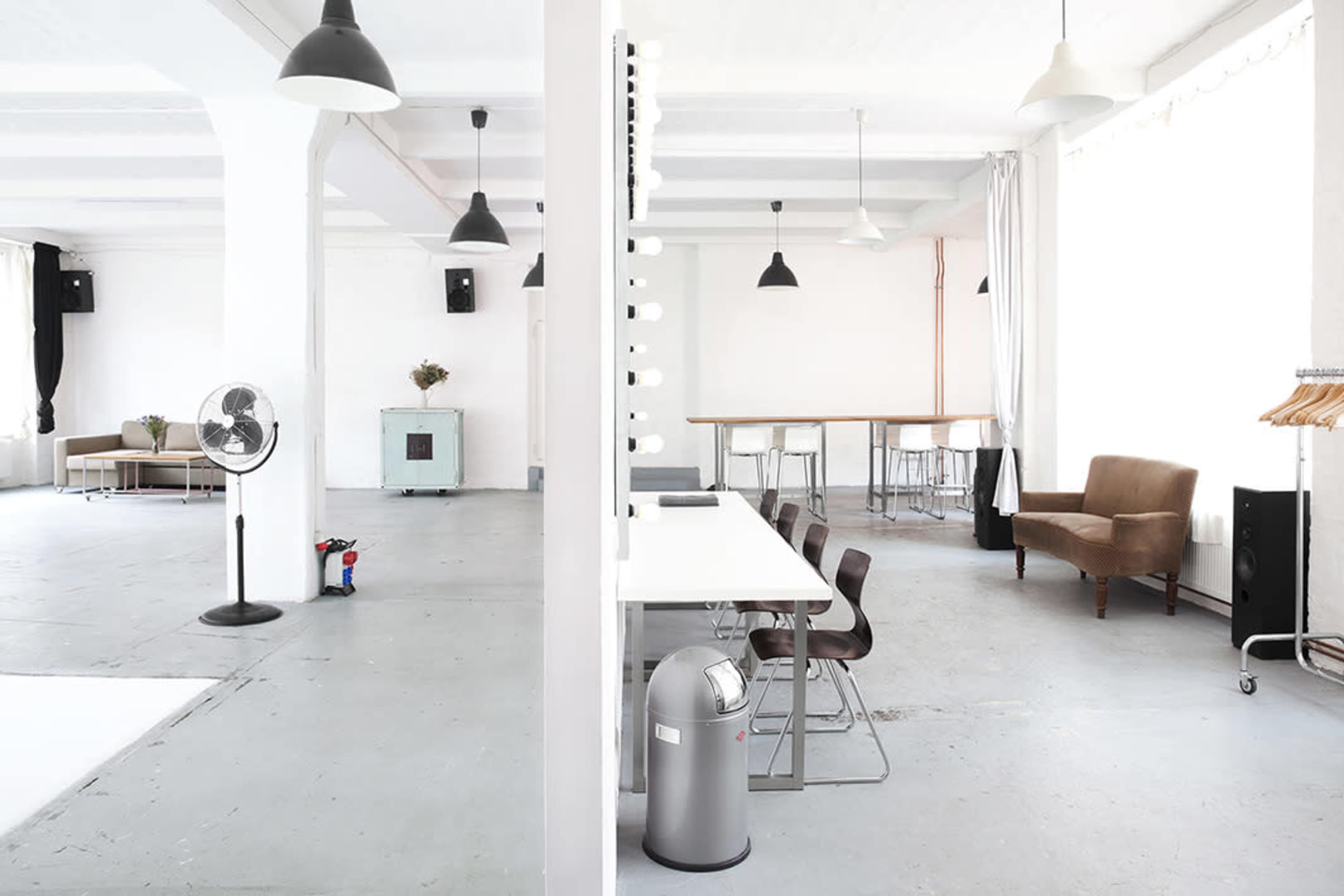A spacious, well-lit studio interior with a mix of seating arrangements, including a brown chair, a row of metal chairs at a table, and a clothing rack.