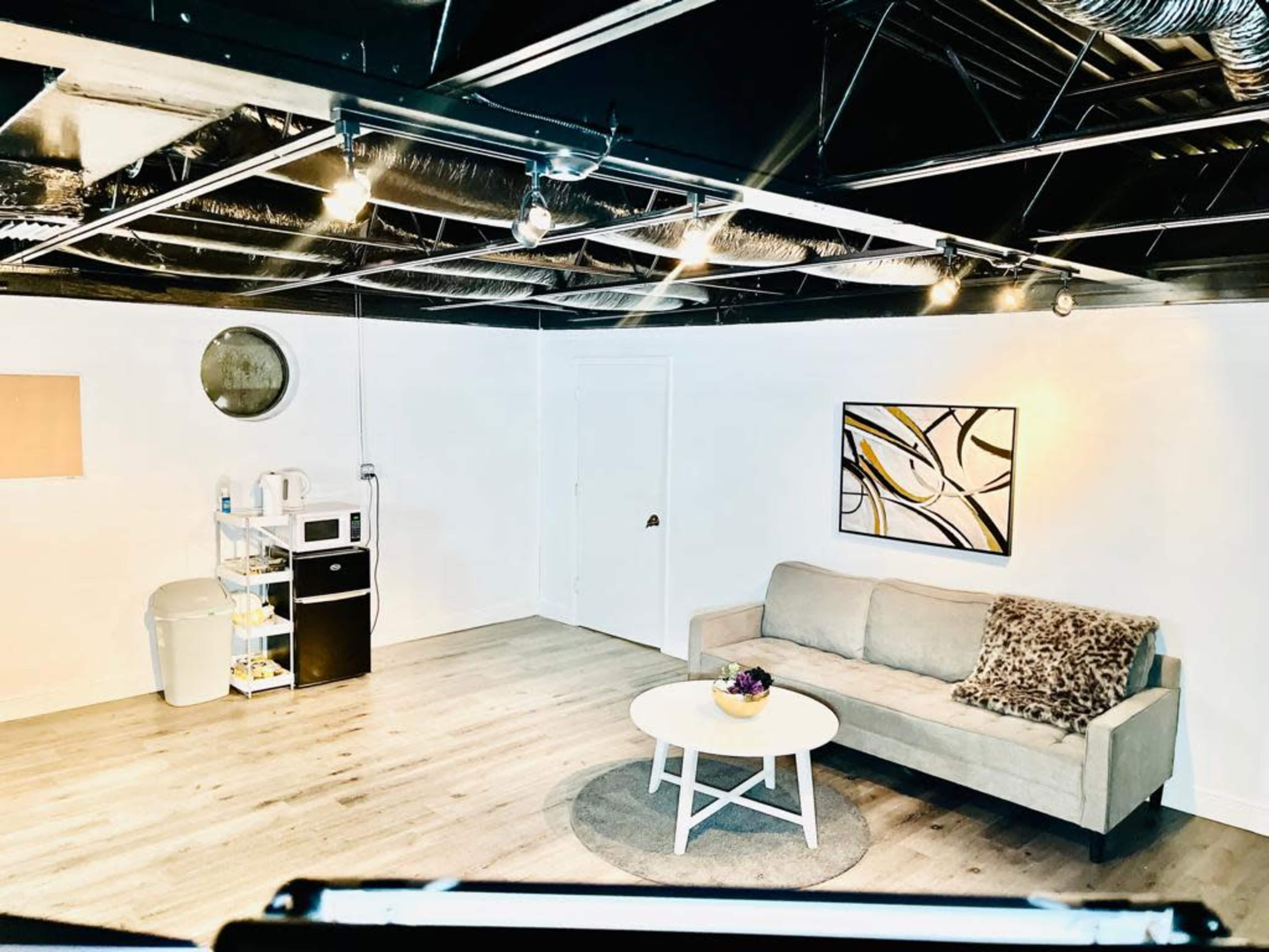 A modern room features a light gray sofa, a round coffee table with a floral centerpiece, a microwave, a small black refrigerator, and a round wall mirror against a white backdrop.