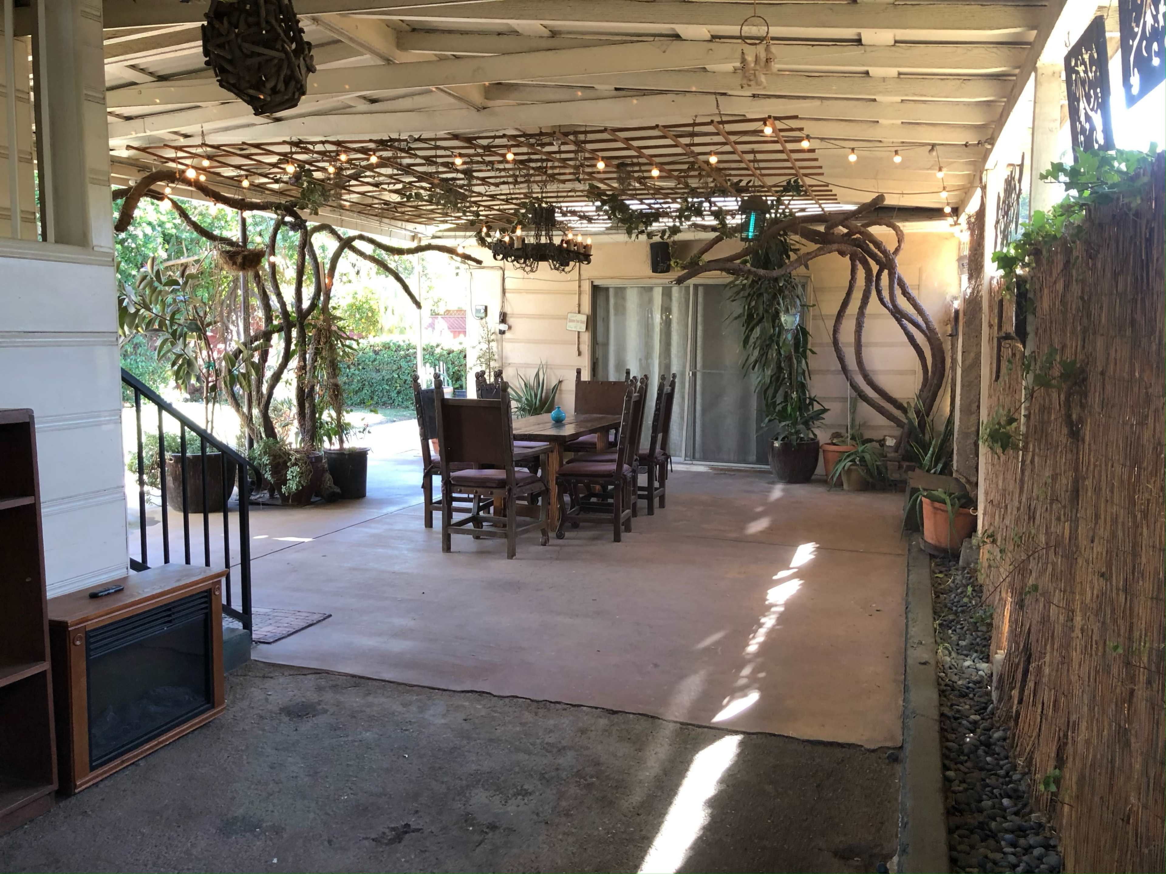 The image shows a covered outdoor patio area with a wooden table and chairs surrounded by greenery and decorative elements.